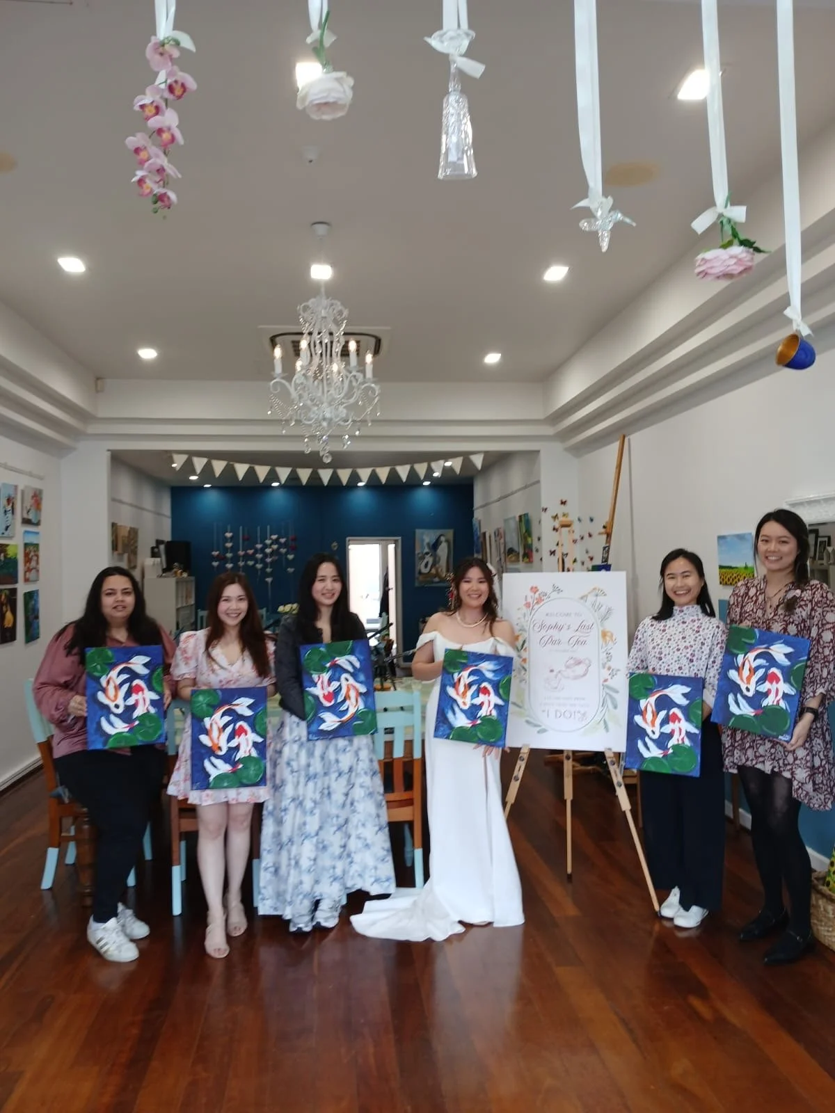 A group of seven women standing indoors in front of a sign and artwork, holding colorful paintings of koi fish on canvases, celebrating an event, with decorations hanging from the ceiling.