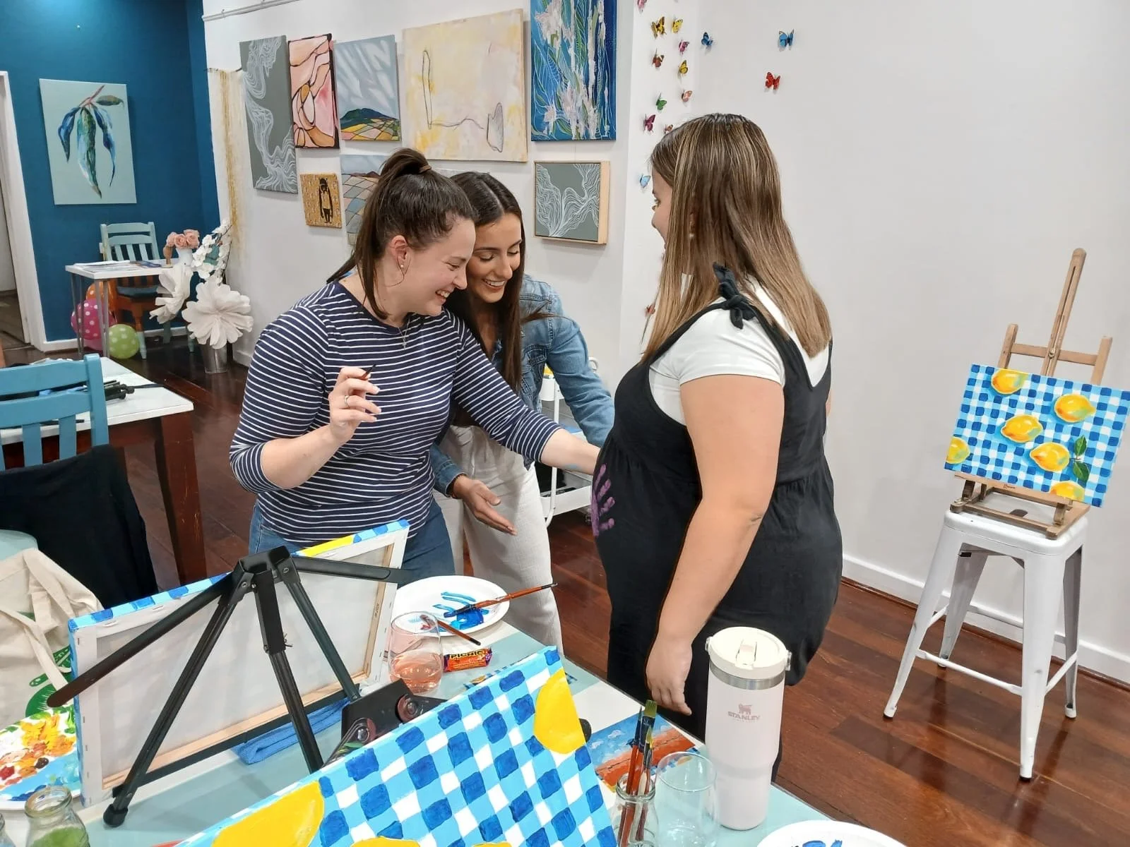 Three women in an art studio laughing and talking, one of them appears to be the artist showing her work. The studio has various paintings on the wall and a small painting of lemons on an easel.