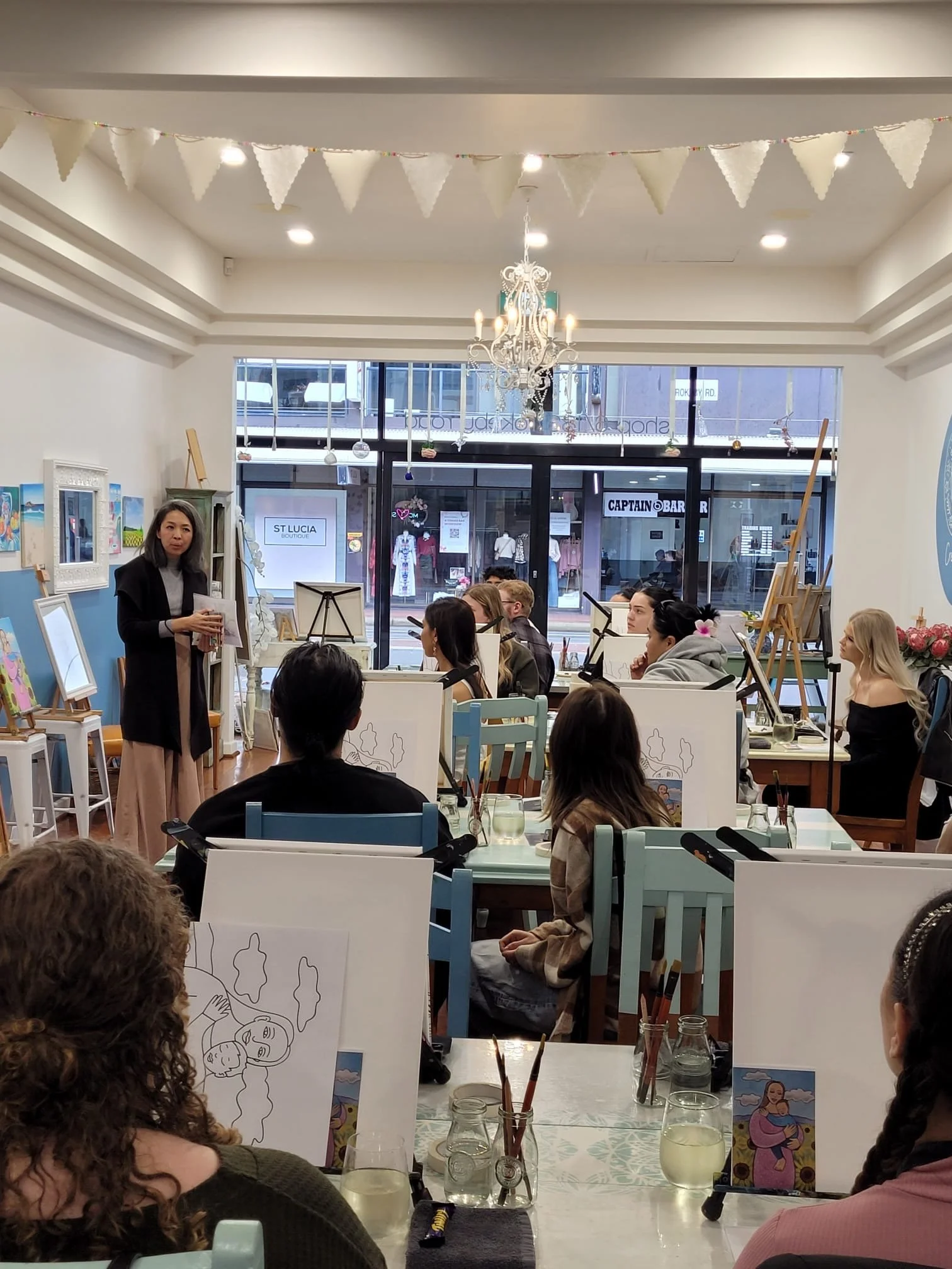 An art class in a bright studio with students painting on easels, a woman instructor standing at the front, and decorated walls with framed artwork, a chandelier, and large windows showing shops outside.