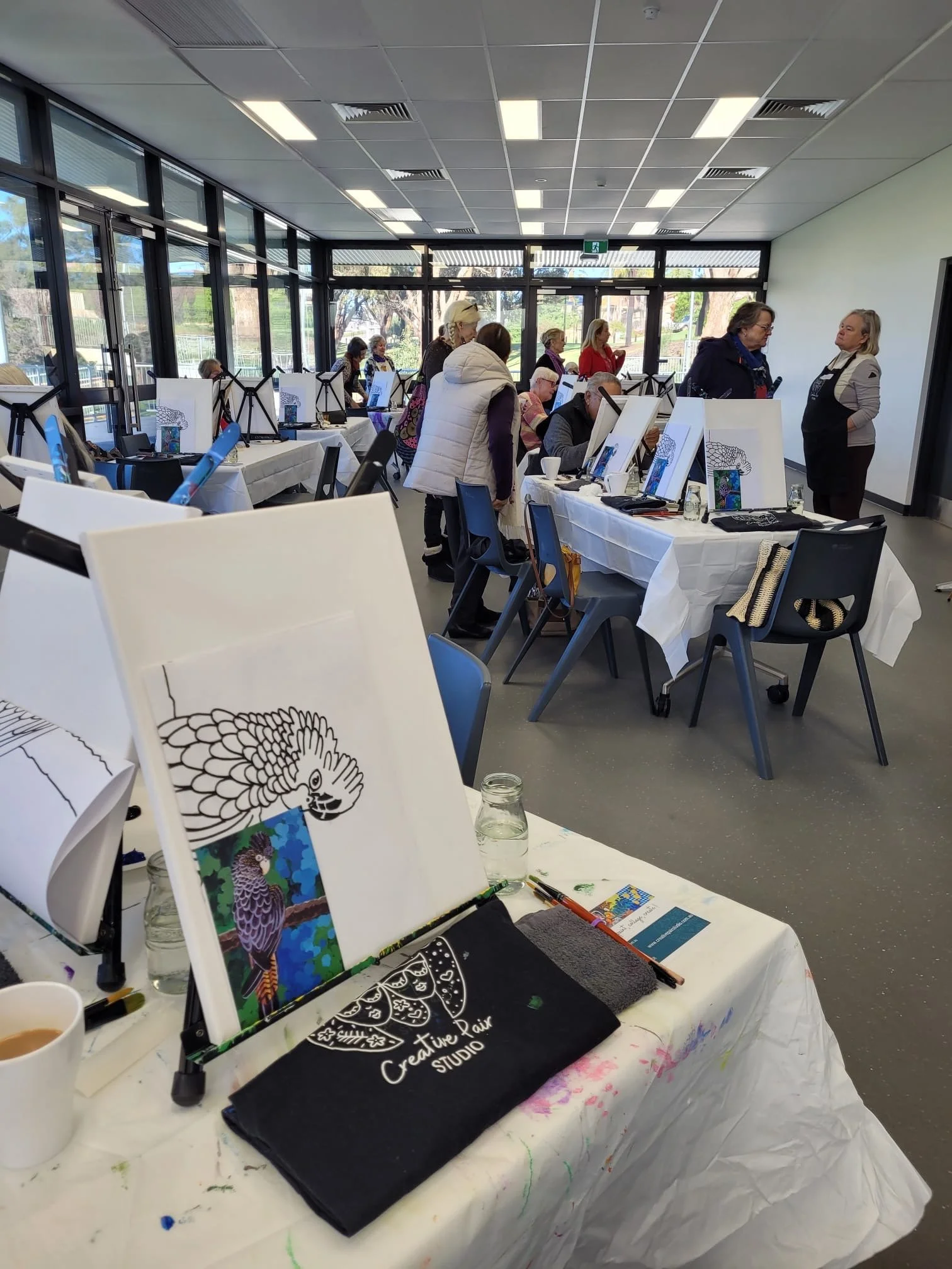 People participating in a painting class in a bright, spacious room with large windows, tables with art supplies, and an instructor guiding students.