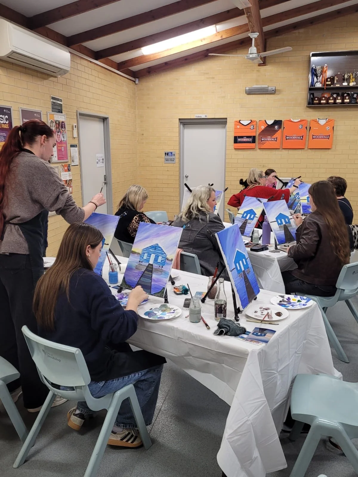 People participating in a painting class, sitting at long tables with canvas paintings of a blue house, in an indoor classroom with yellow brick walls.