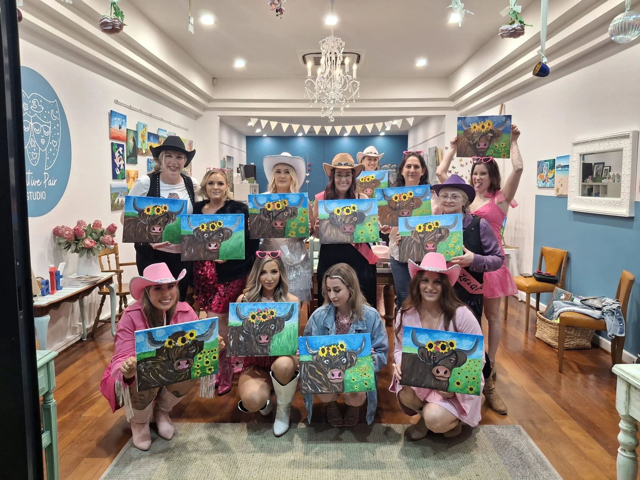 Group of women in a studio, some wearing cowboy hats, holding paintings of a buffalo with a sunflower crown in bright colors. They are smiling in a decorated room with a chandelier, floral arrangements, and various artwork on the walls.