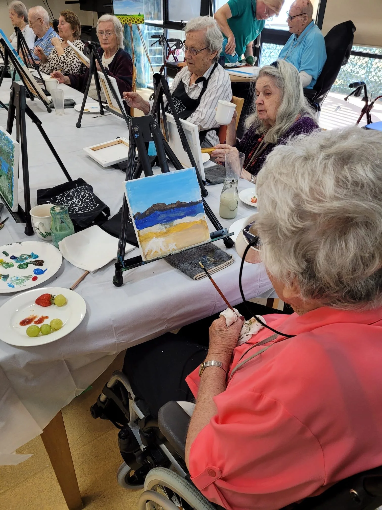 A group of elderly women participating in a painting class, seated at a long table with easels, paints, and brushes. One woman in a wheelchair is painting with assistance, and finished landscape paintings are visible on their canvases. Some women are holding drinks and snacks.