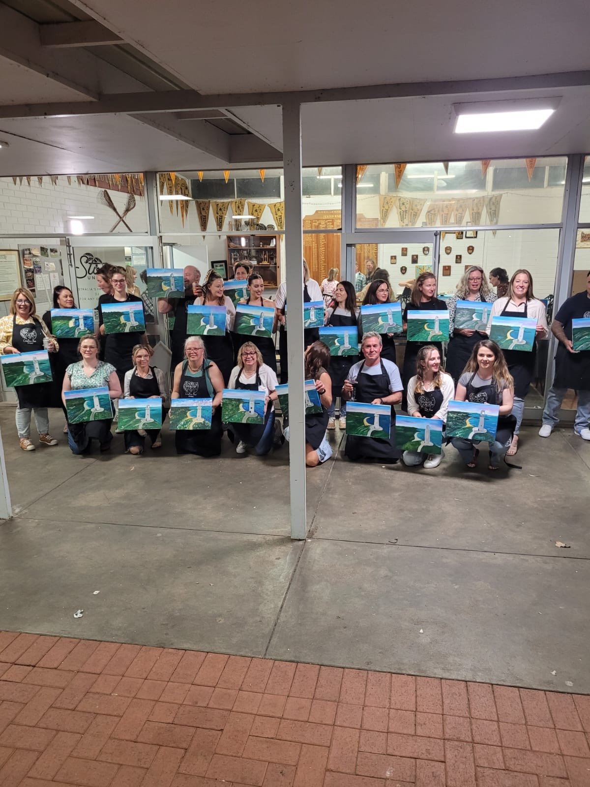 Group of people participating in a painting event, holding their completed landscape paintings of waterfalls and mountains, indoors in a well-lit space with large windows and decorative banners.