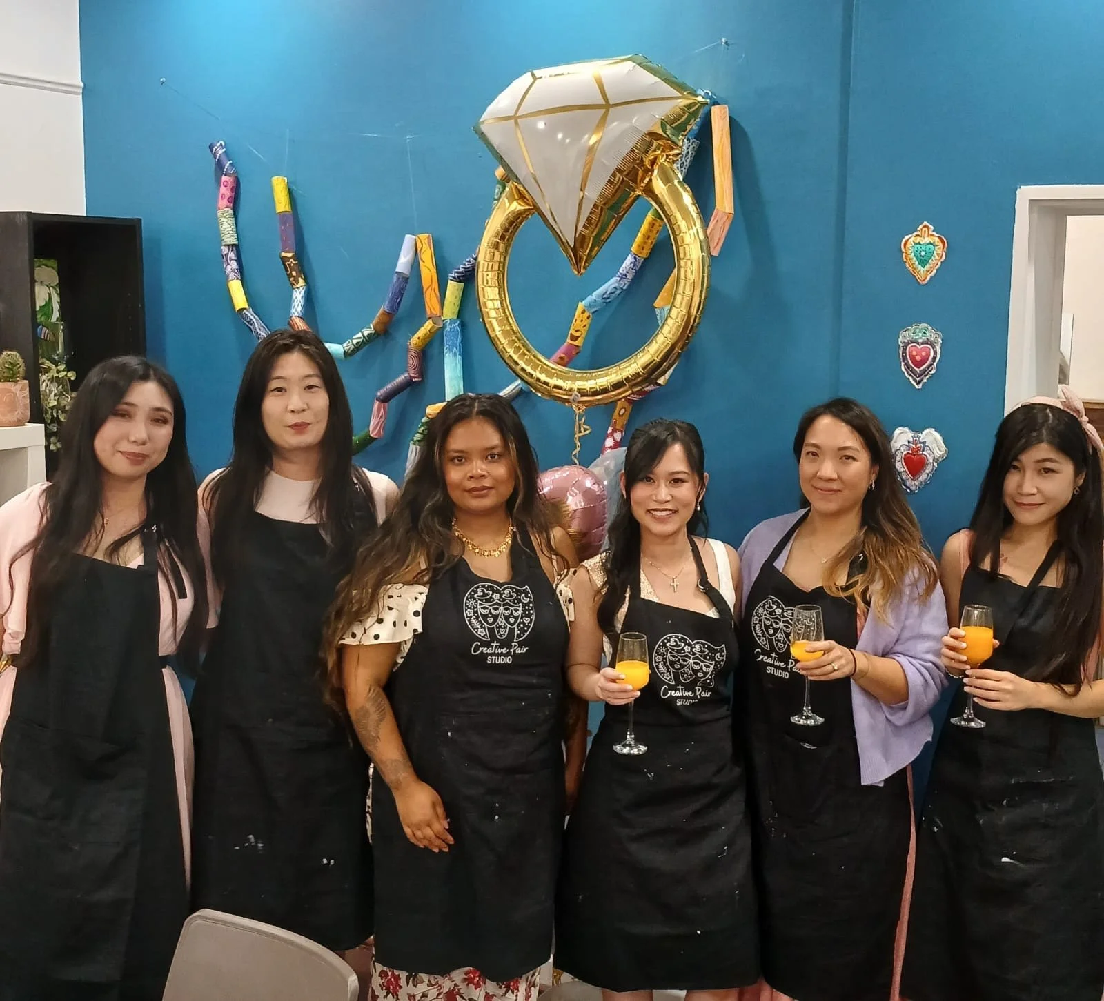 Group of six women standing together at a celebration, wearing aprons, holding glasses with orange juice, and smiling, with colorful balloons and decorations on the blue wall behind them.