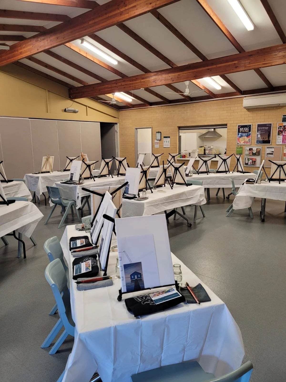A room set up for an event with several tables covered in white tablecloths, each with a music stand, small electronic devices, and informational materials. The room has a high ceiling with wooden beams, fluorescent lighting, and a kitchen area in the background.