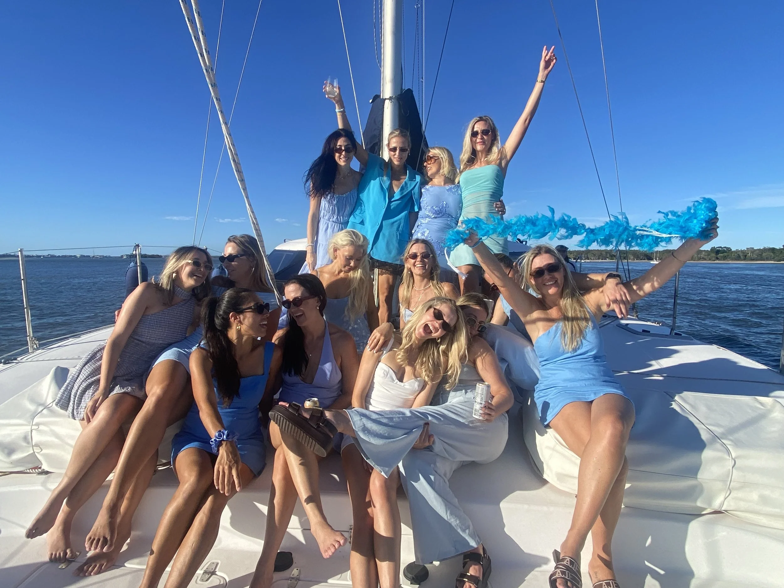 Group of women enjoying a boat ride on a sunny day, some wearing blue dresses, with blue sky and water in the background.