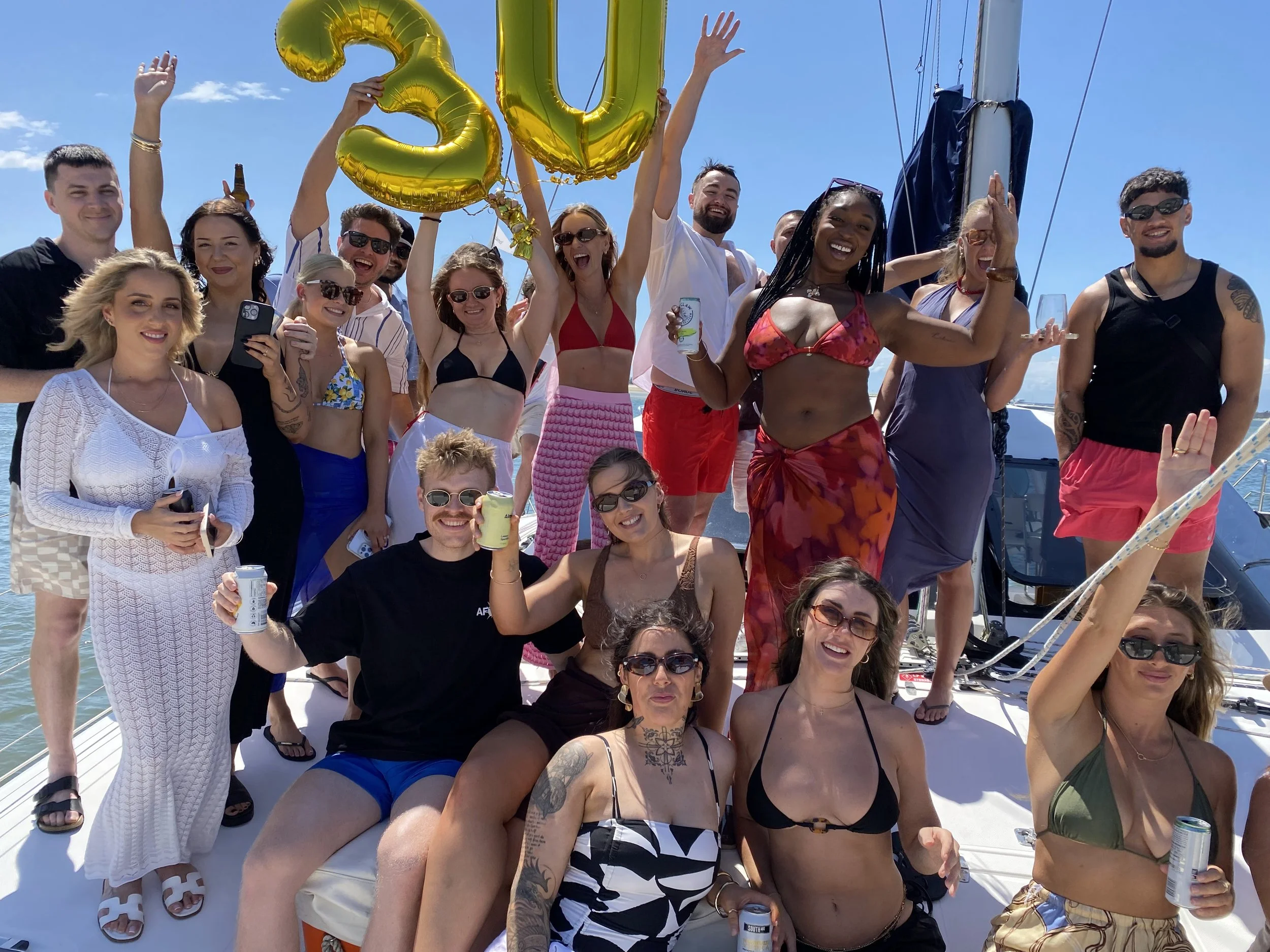 Group of people celebrating on a boat under blue sky, holding gold balloons forming the number 30.