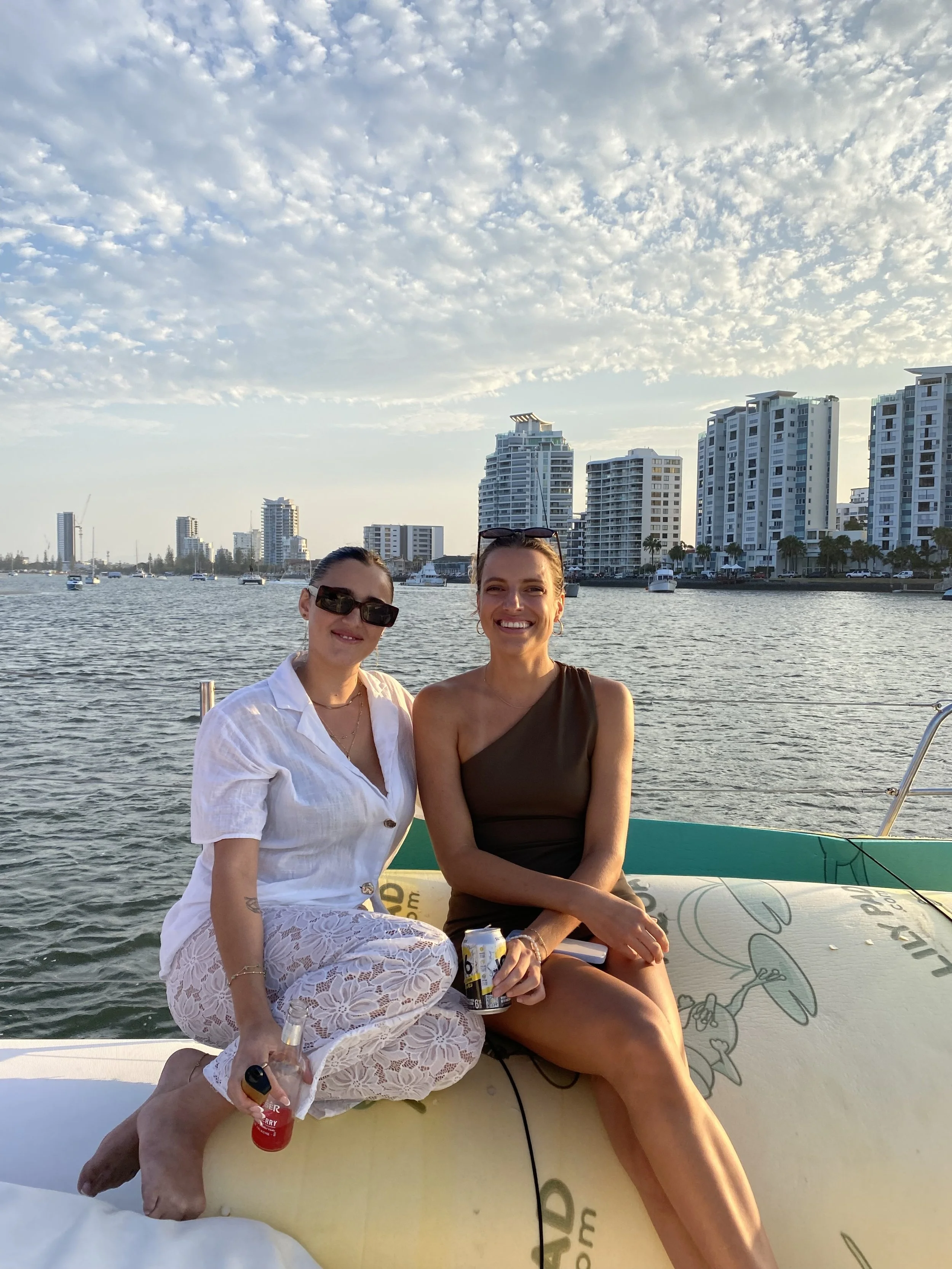 Two women sitting on a paddleboard in a marina, with city high-rise buildings and a partly cloudy sky in the background, enjoying a sunny day.