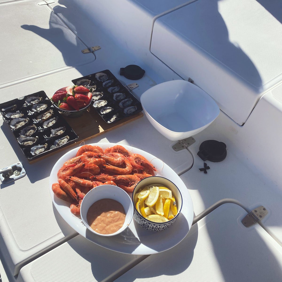 Seafood on a boat deck including a plate of cooked shrimp, a bowl of lemon slices, a bowl of sauce, and trays of oysters and strawberries.