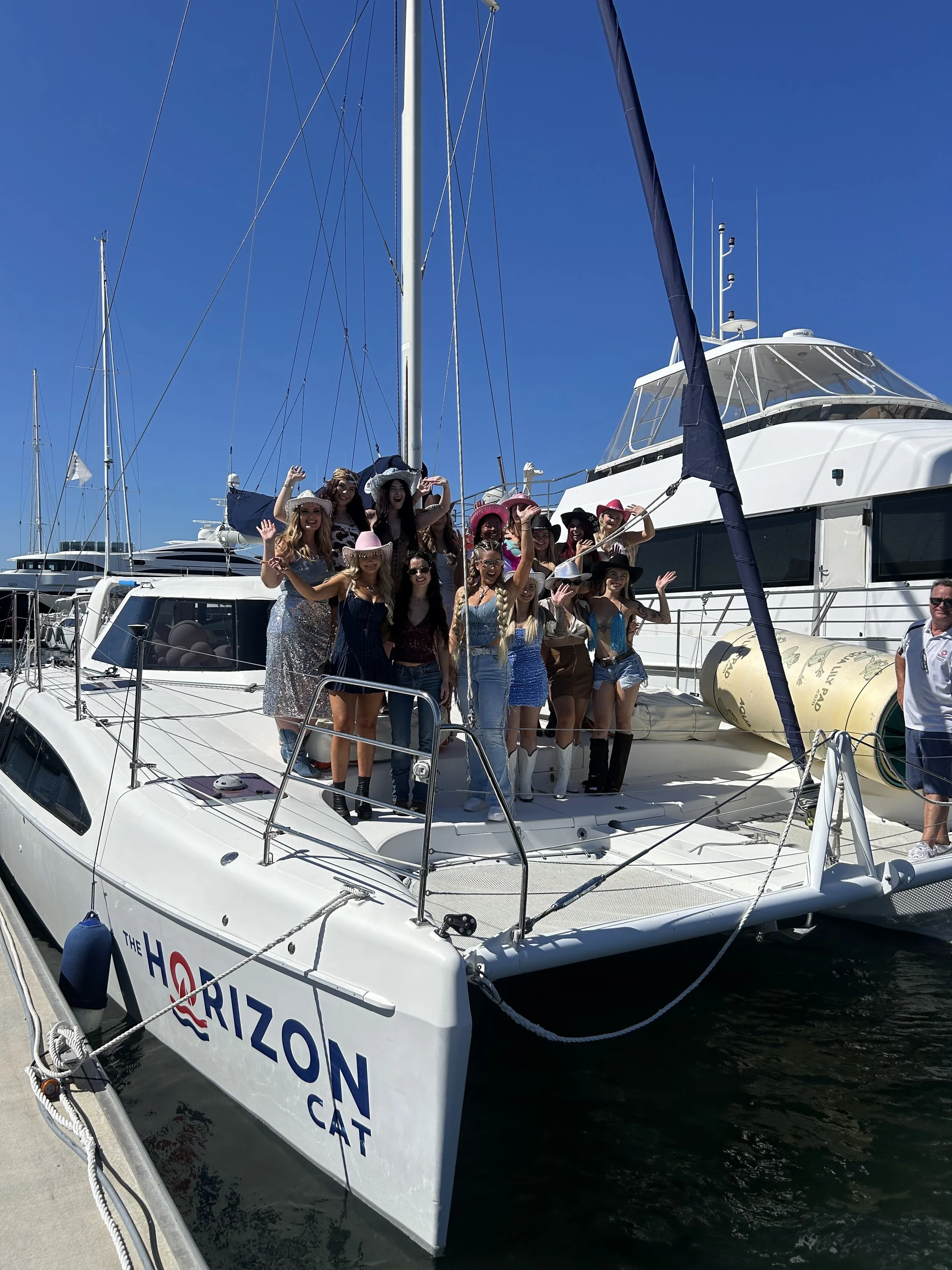 A group of women in fashionable outfits and hats standing on a boat at a marina, waving and smiling with other boats in the background and a clear blue sky.
