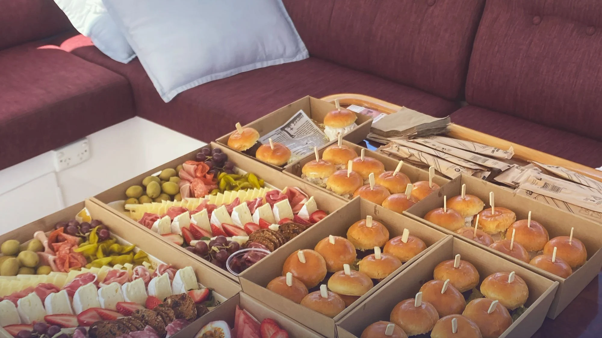 A table with multiple boxes of assorted sandwiches, mini burgers, chocolates, fruits, and napkins in a living room setting.