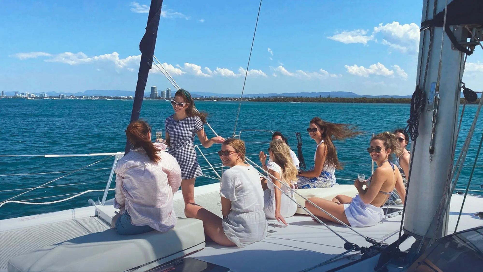 Group of women enjoying a boat ride on a sunny day with blue water and city skyline in the background.