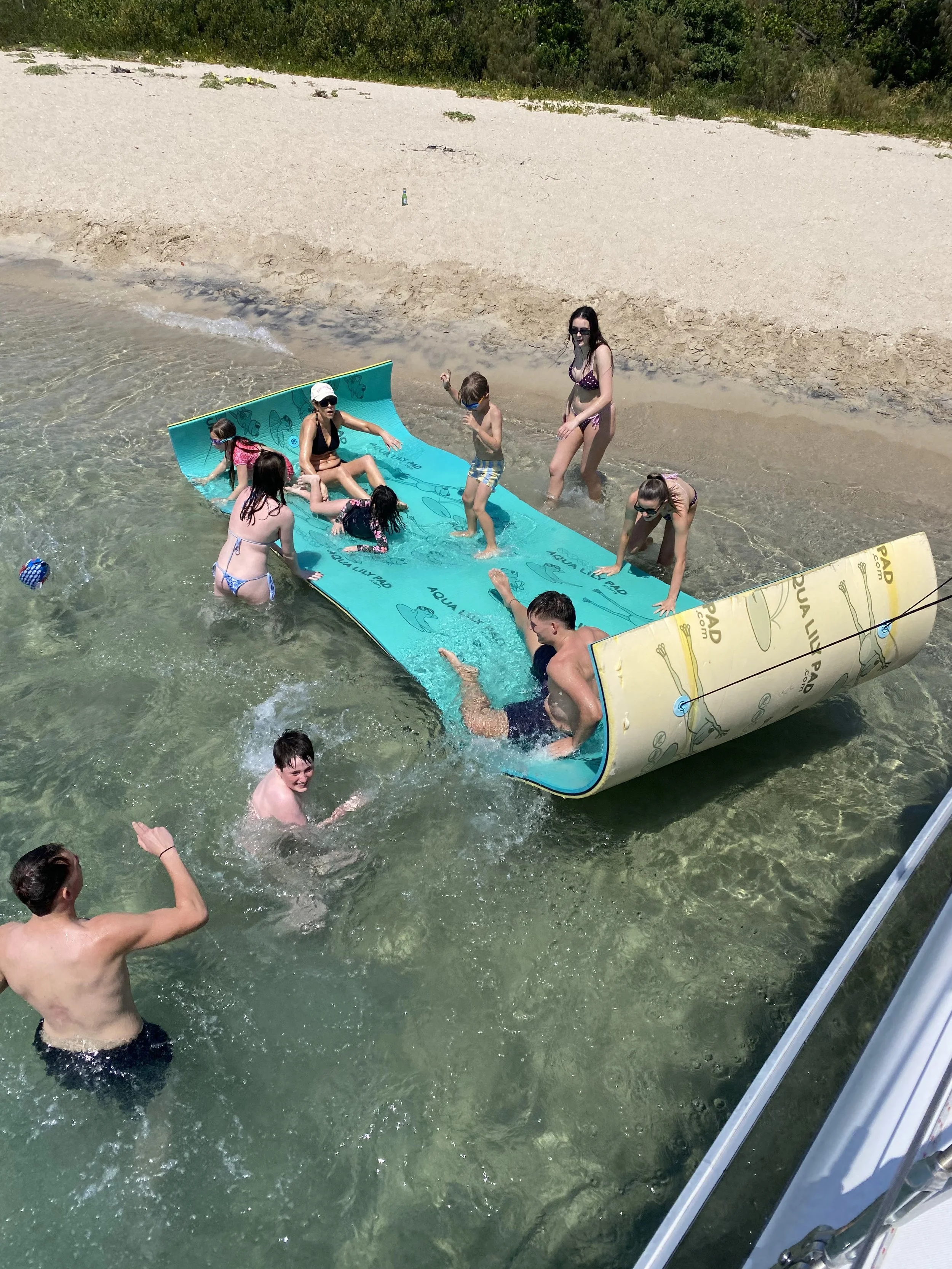 Group of children and adults playing on an overturned inflatable raft in shallow water near a sandy beach on a sunny day.