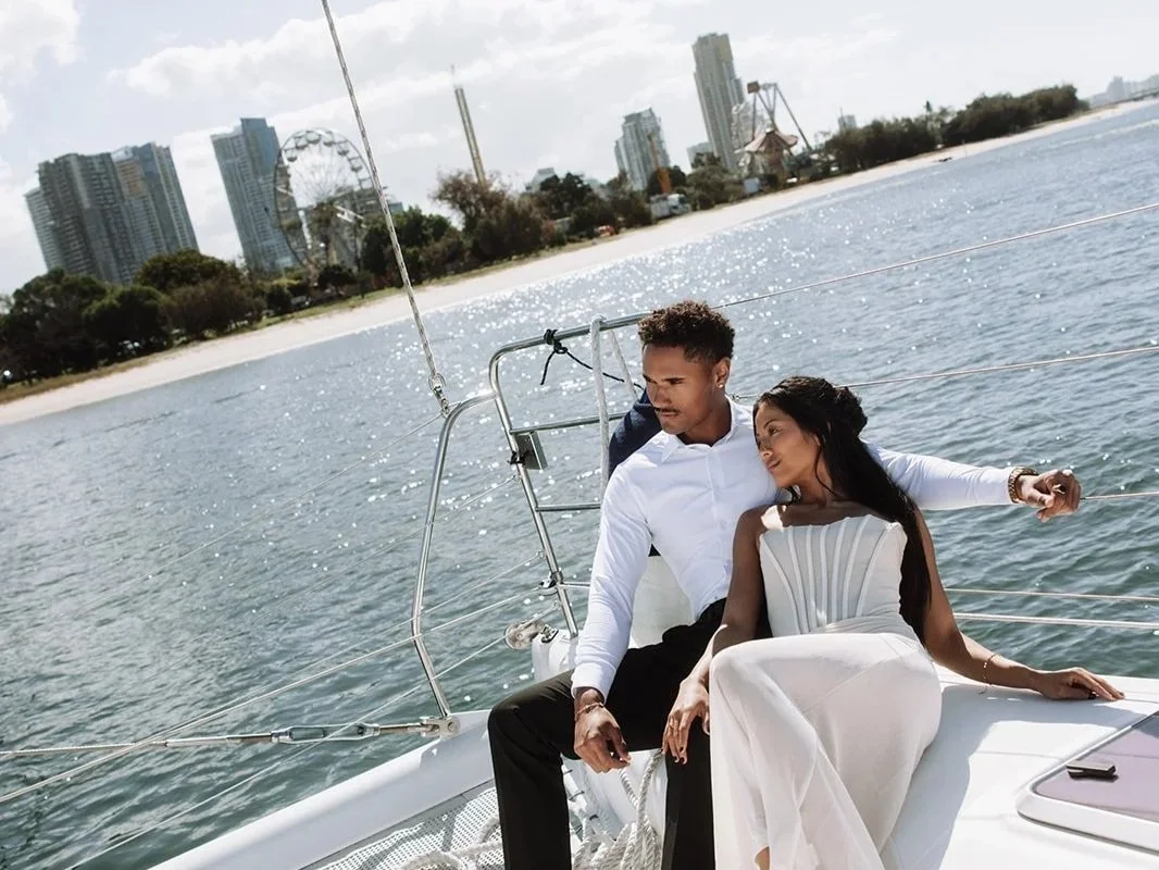 A couple sitting on a yacht, with a city skyline and a Ferris wheel in the background, enjoying a sunny day on the water.