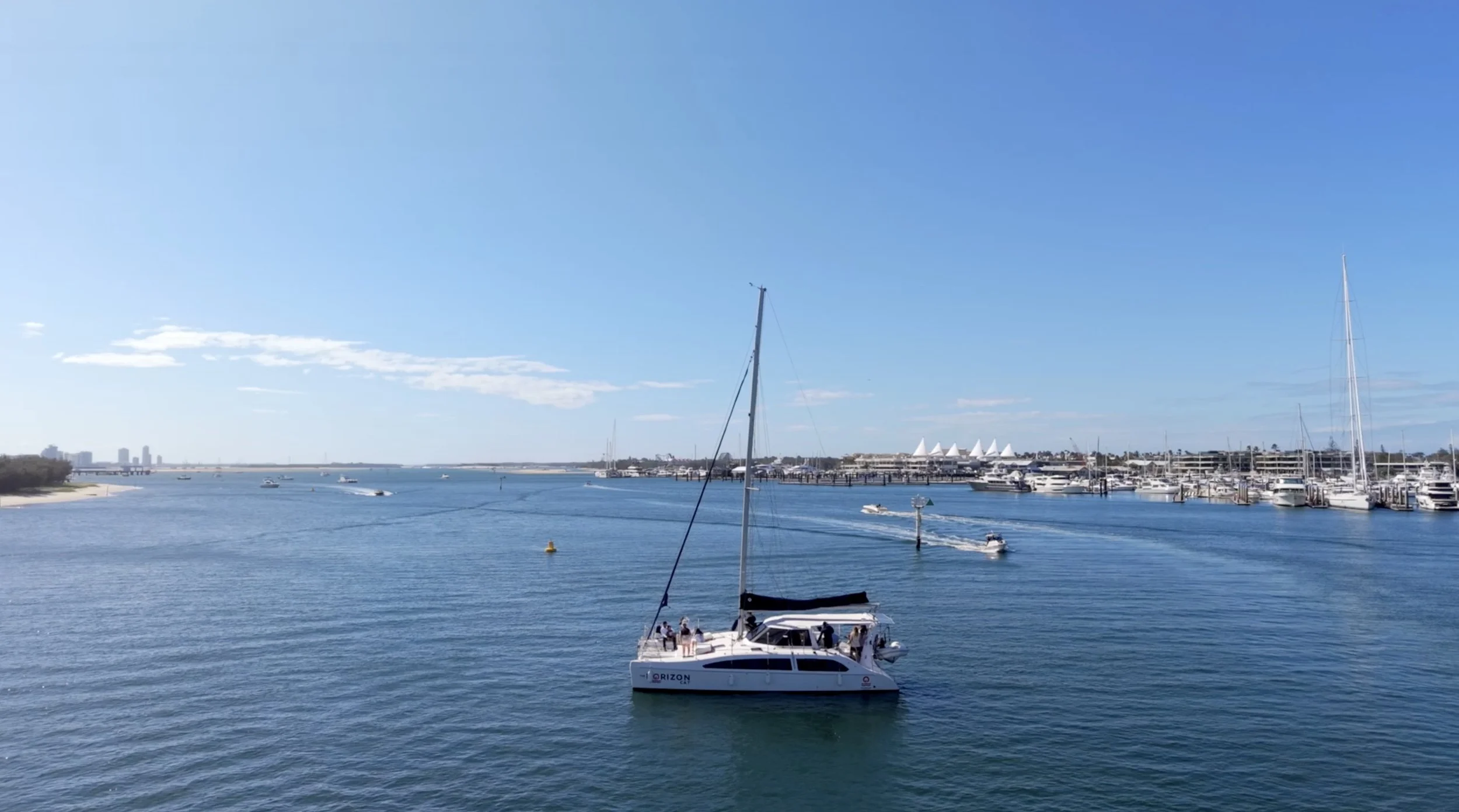 Sailboats and yachts moored in a marina under a clear blue sky, with city buildings in the distance.