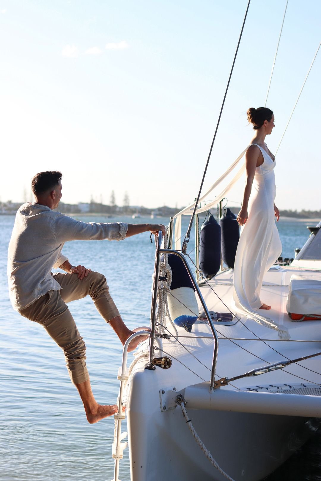 A man climbing onto a sailboat while a woman stands on the deck, with water and a distant shoreline in the background.