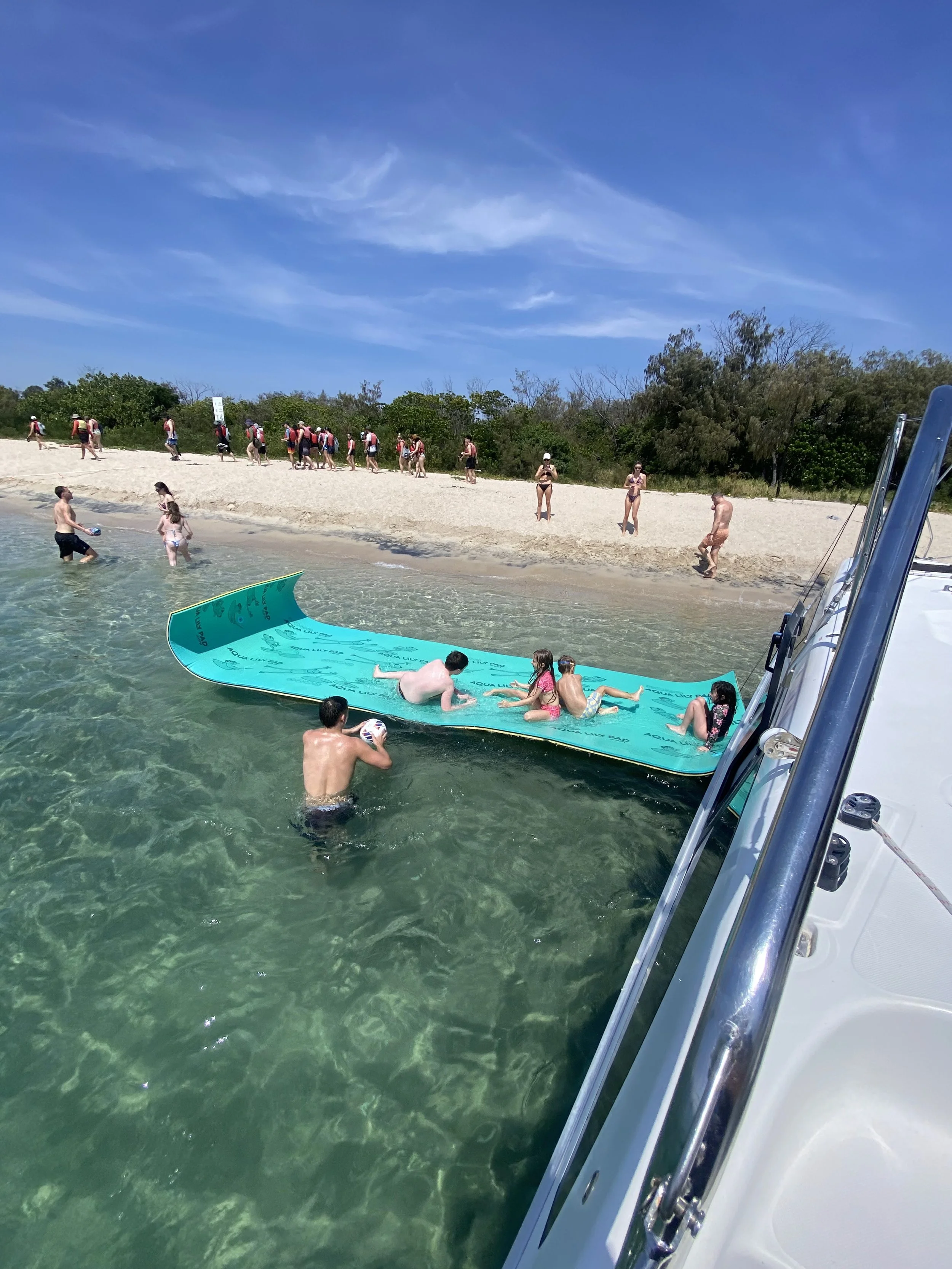 People swimming and lounging on a floating mat near the shoreline of a beach with a sandy shore, green trees, and a bright blue sky with some clouds.
