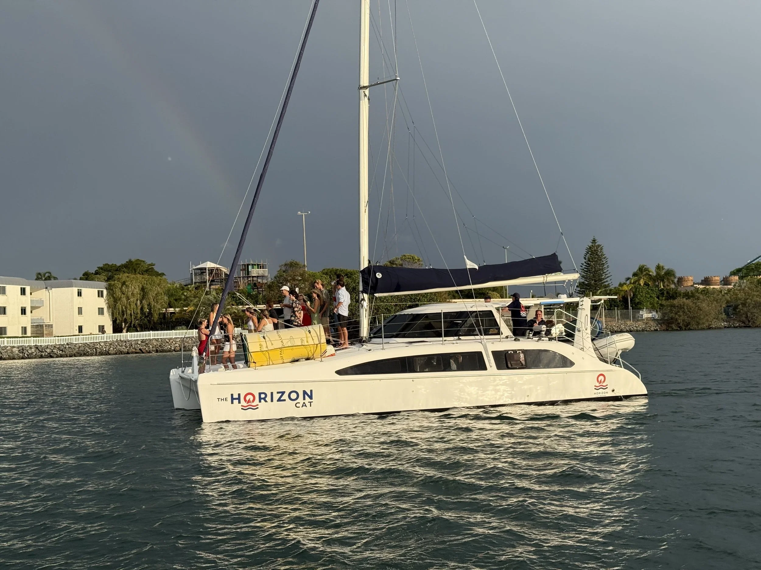 A white yacht with the words 'The Horizon Cat' on the side is on the water with several people onboard. The sky is partly cloudy, and there is a faint rainbow in the background.