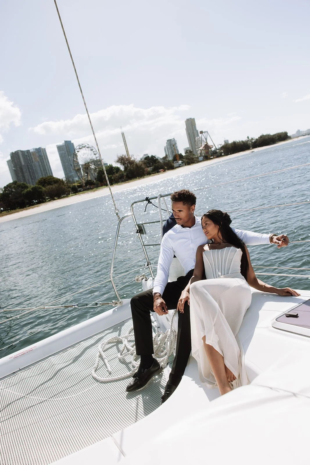 A couple sitting close together on a boat, with a city skyline and amusement park in the background, enjoying a sunny day on the water.