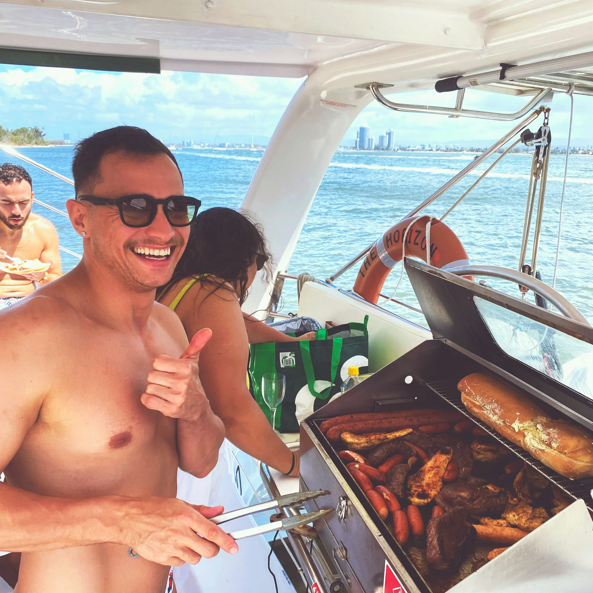 Man smiling and giving a thumbs-up while grilling sausages and meats on a boat. Two other people are in the background, with a city skyline and water view behind them.