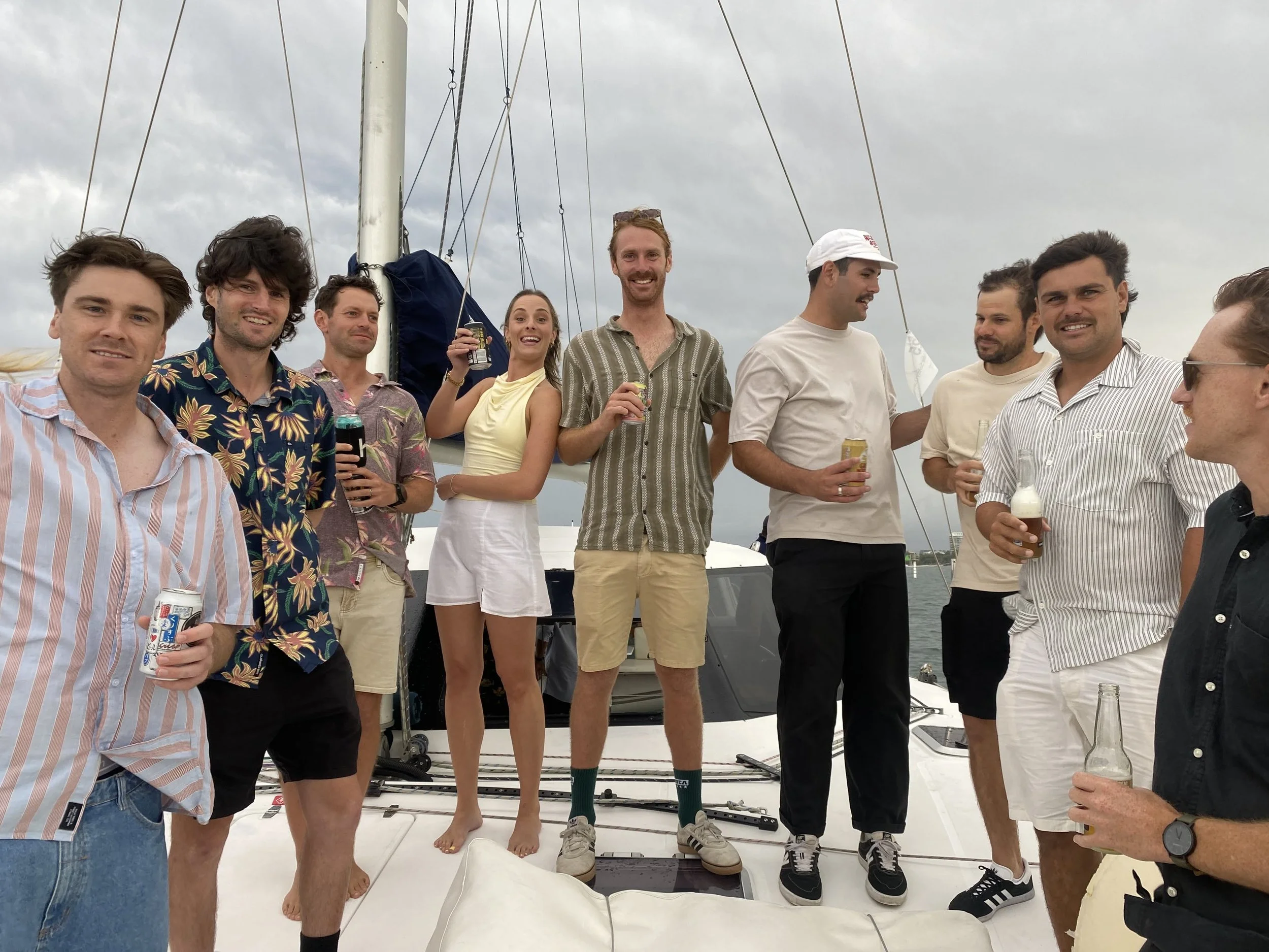 A group of nine people on a boat, holding drinks, smiling, in casual summer clothing, with a cloudy sky in the background.