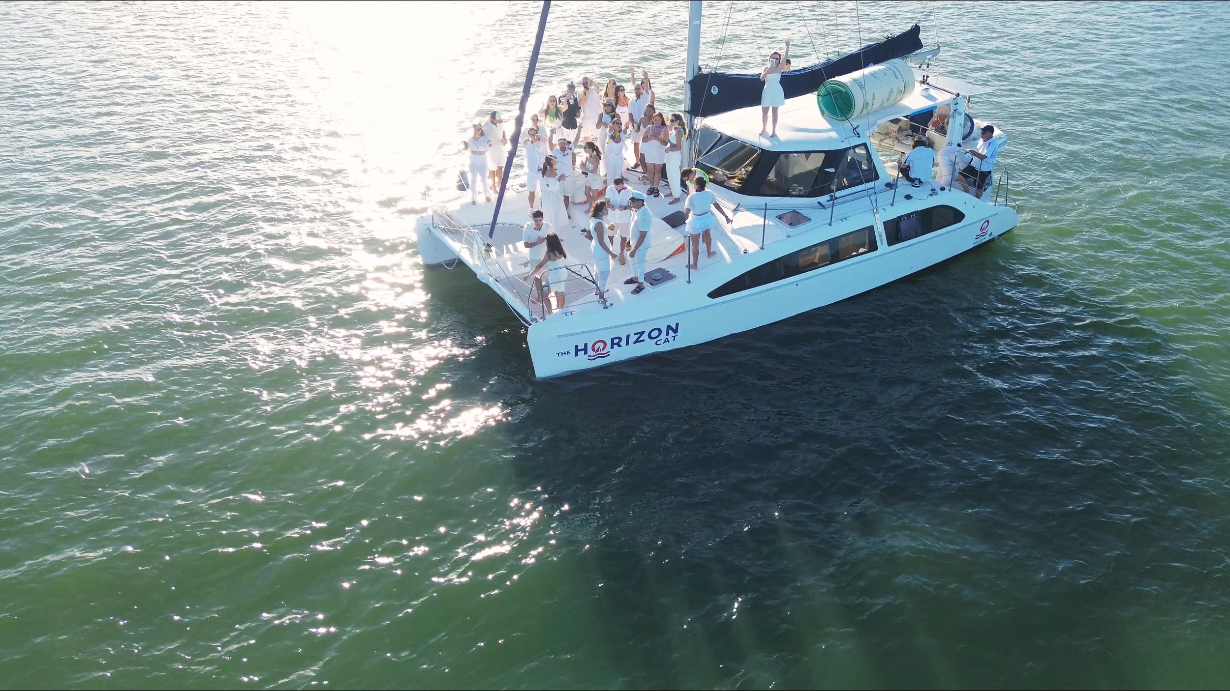 A large group of people dressed in white on a white yacht named 'The Horizon Cat' sailing on the water. Some are standing, some are sitting, and a few are dancing or waving. The boat is in bright sunlight, creating reflections on the water.