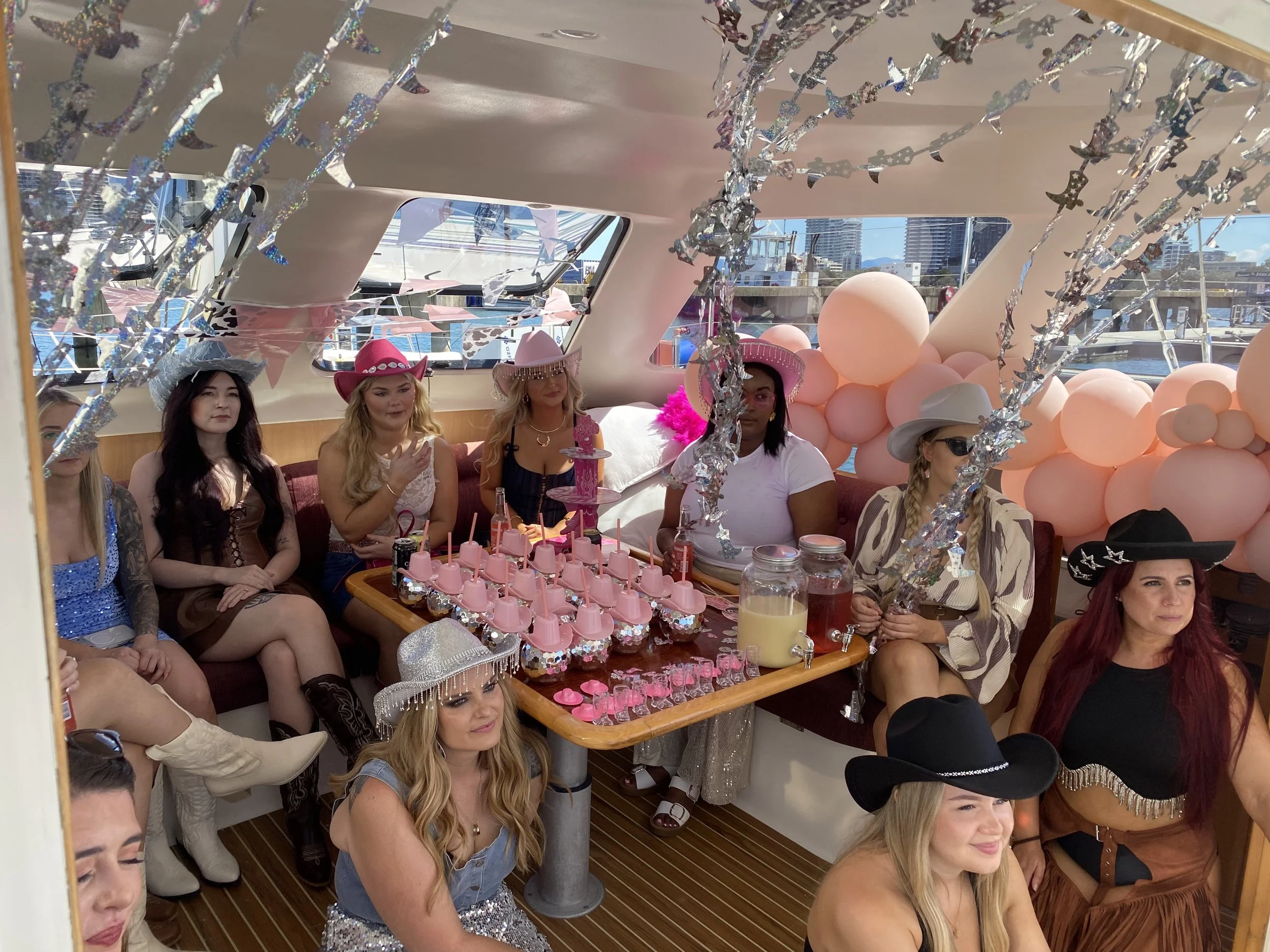 Women dressed in cowboy and western-themed party costumes sitting inside a yacht decorated with pink and silver balloons and streamers, celebrating a party.