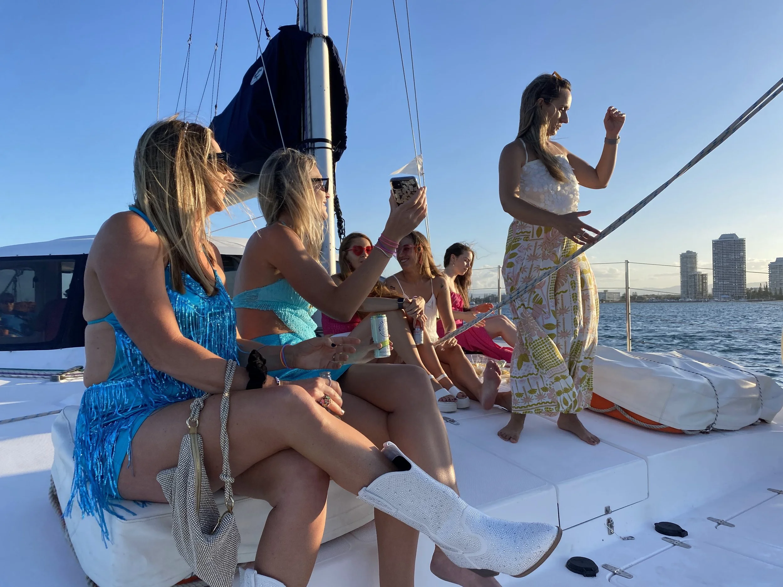 Group of women on a yacht during sunset, some sitting and some standing, enjoying a boat party with city skyline in the background.