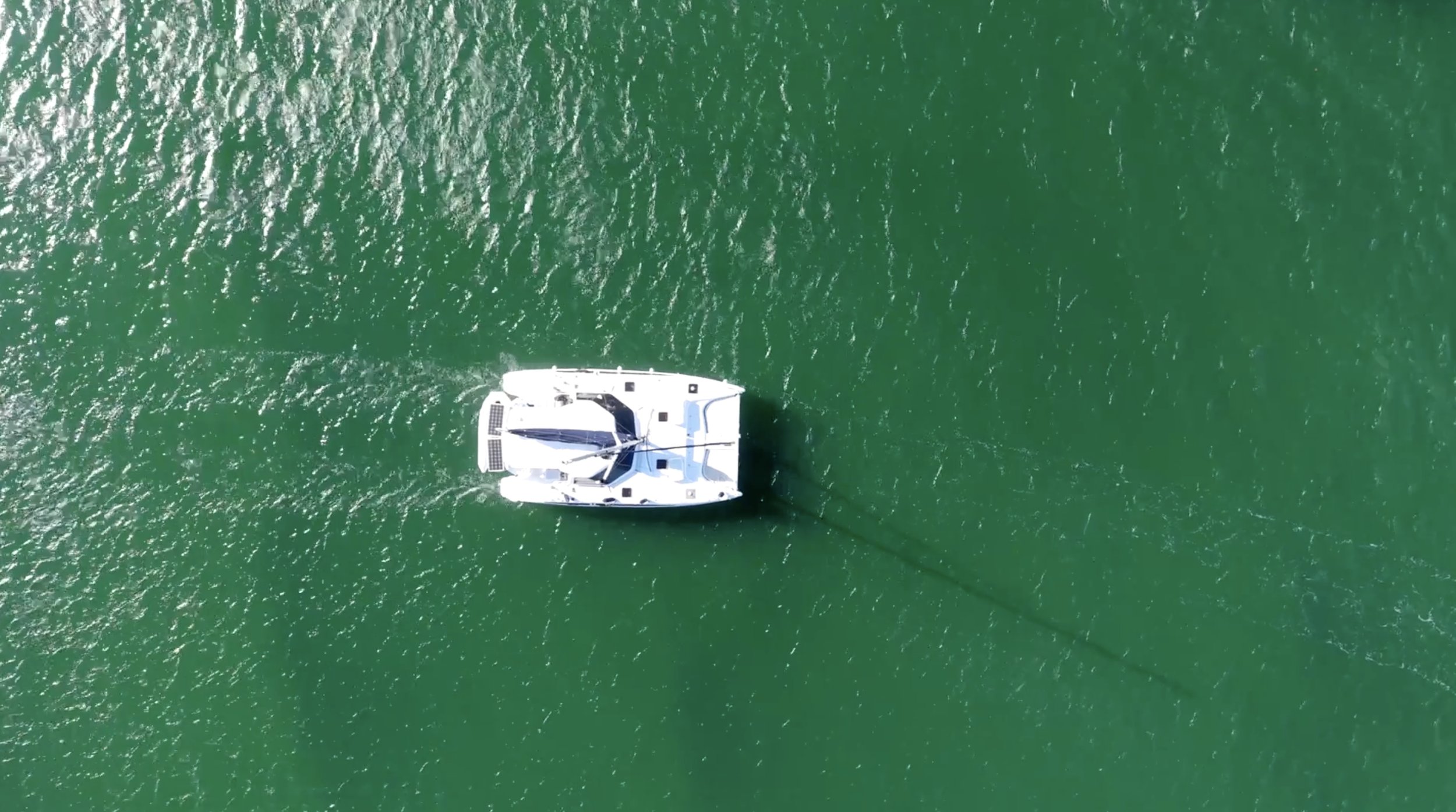 An aerial view of a white sailboat on green water, seen from above.