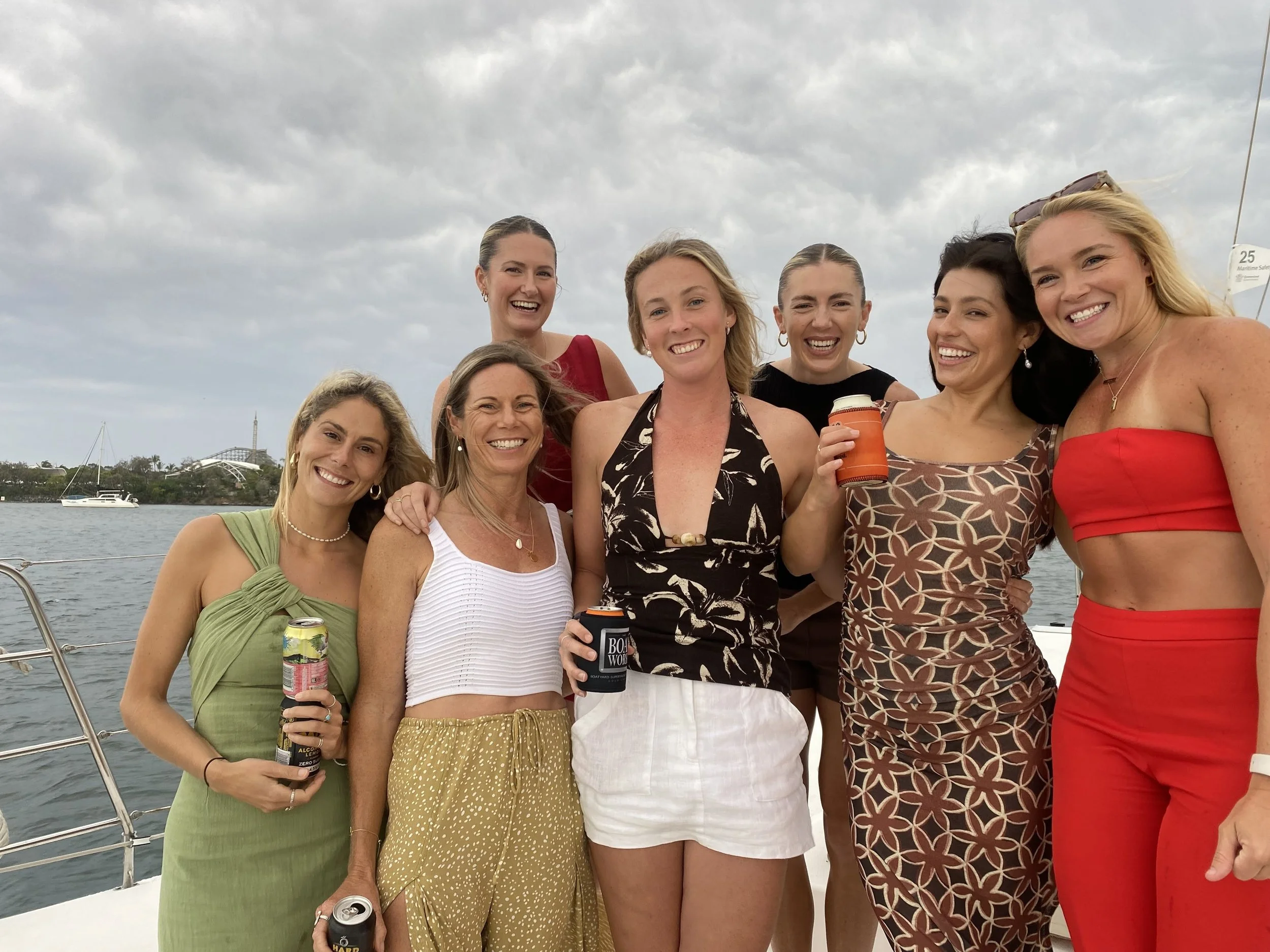 Group of eight women smiling and posing together on a boat, some holding drinks, with water and boats in the background under cloudy skies.