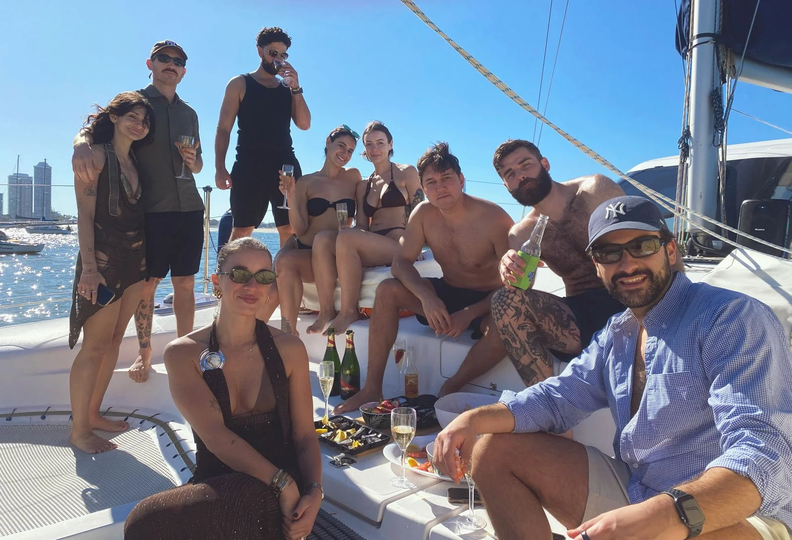 Group of friends on a boat enjoying drinks and snacks on a sunny day with a city skyline in the background.
