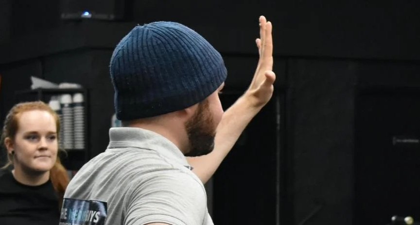 A man in a blue knit beanie and gray shirt raising his hand, standing in front of a woman with red hair, in a room with dark walls.