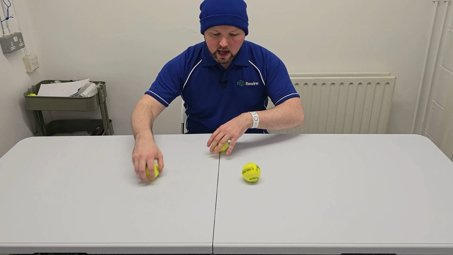 A man wearing a blue shirt and beanie sitting at a table, rolling 3 tennis balls with letters written on them.
