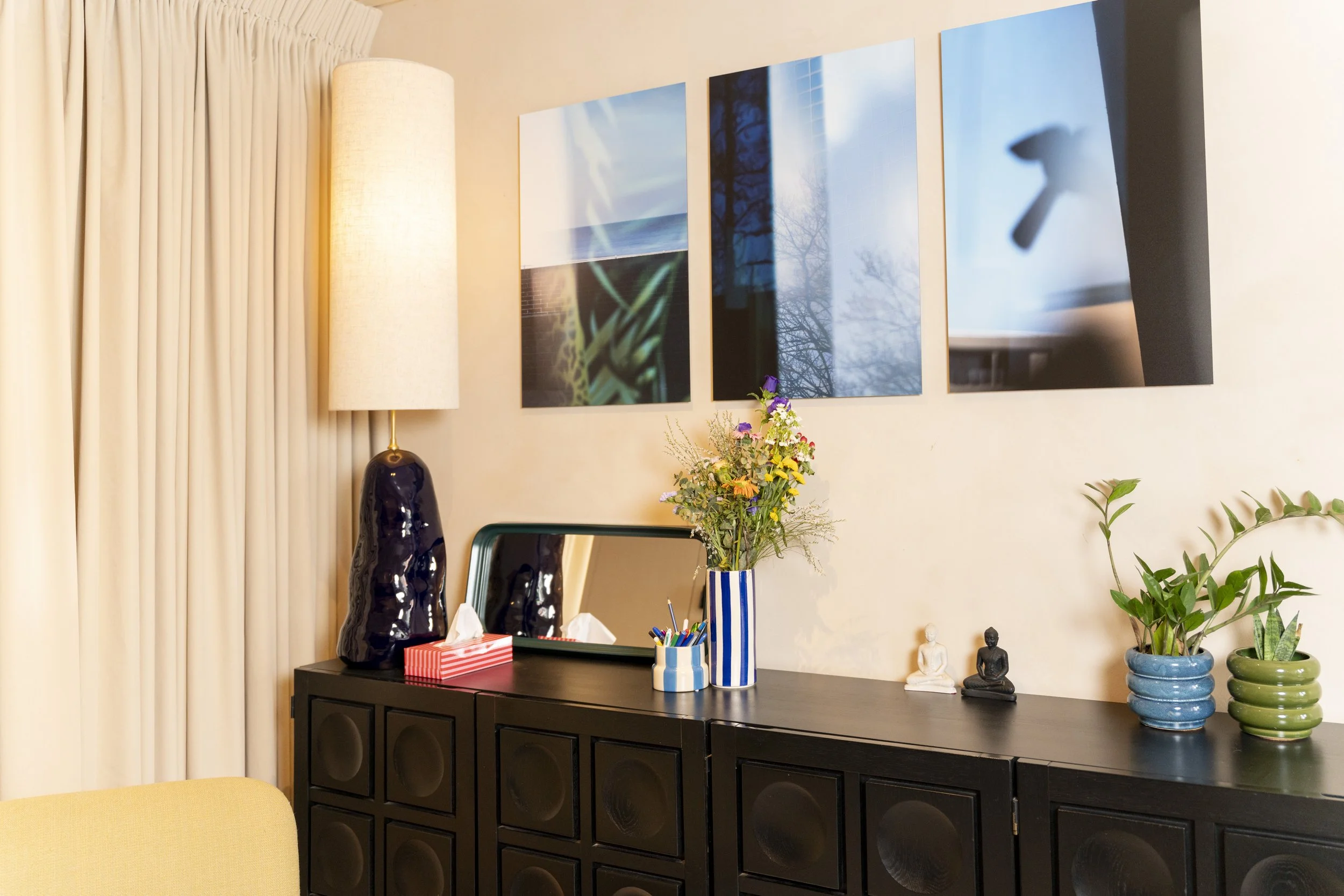 Interieur van een woonkamer met een zwarte dressoir, decoratieve vazen, planten, een spiegel, een lamp en vier kunstwerken aan de muur.