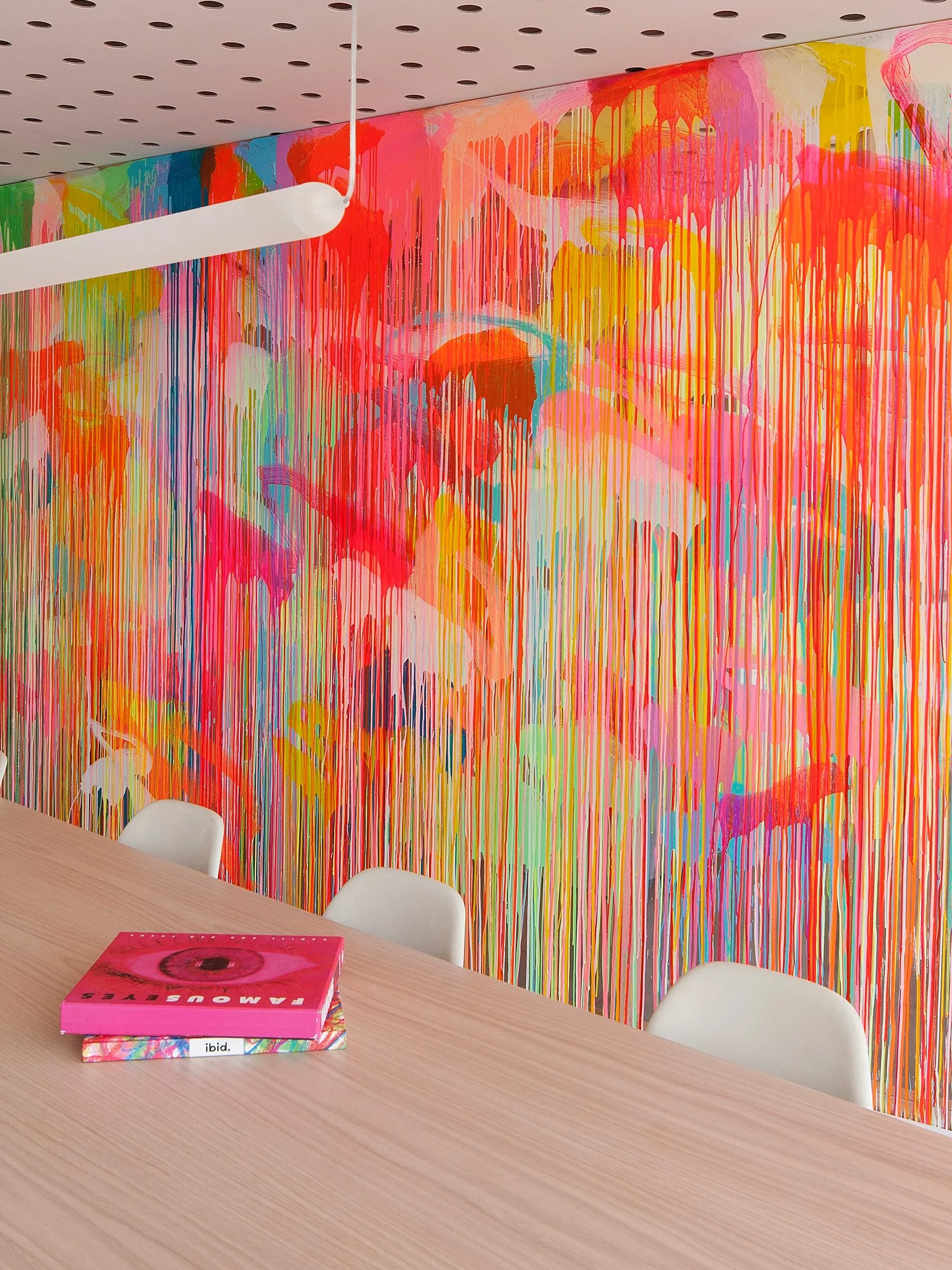 A colorful abstract mural with dripped paint in pink, orange, yellow, green, blue, and purple on a wall. In the foreground, a wooden table with a pink box labeled 'tamouseyes' and a white chair.