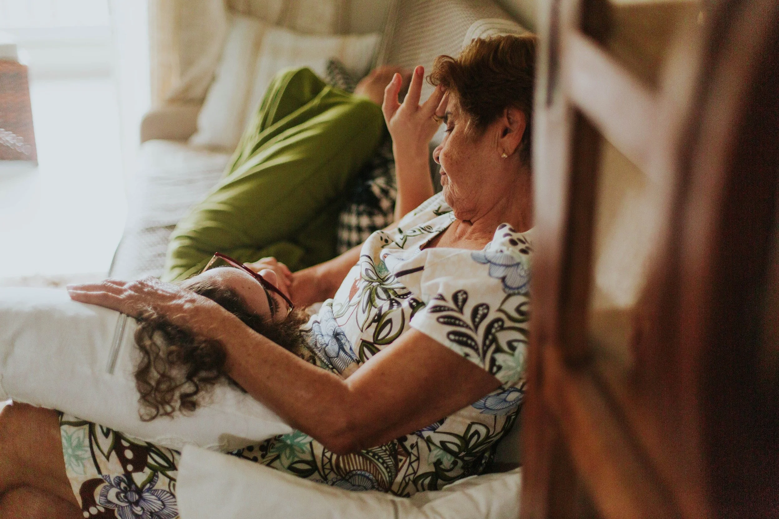 An older woman resting her head in her hand while a younger woman lies beside her, both looking pensive in soft light