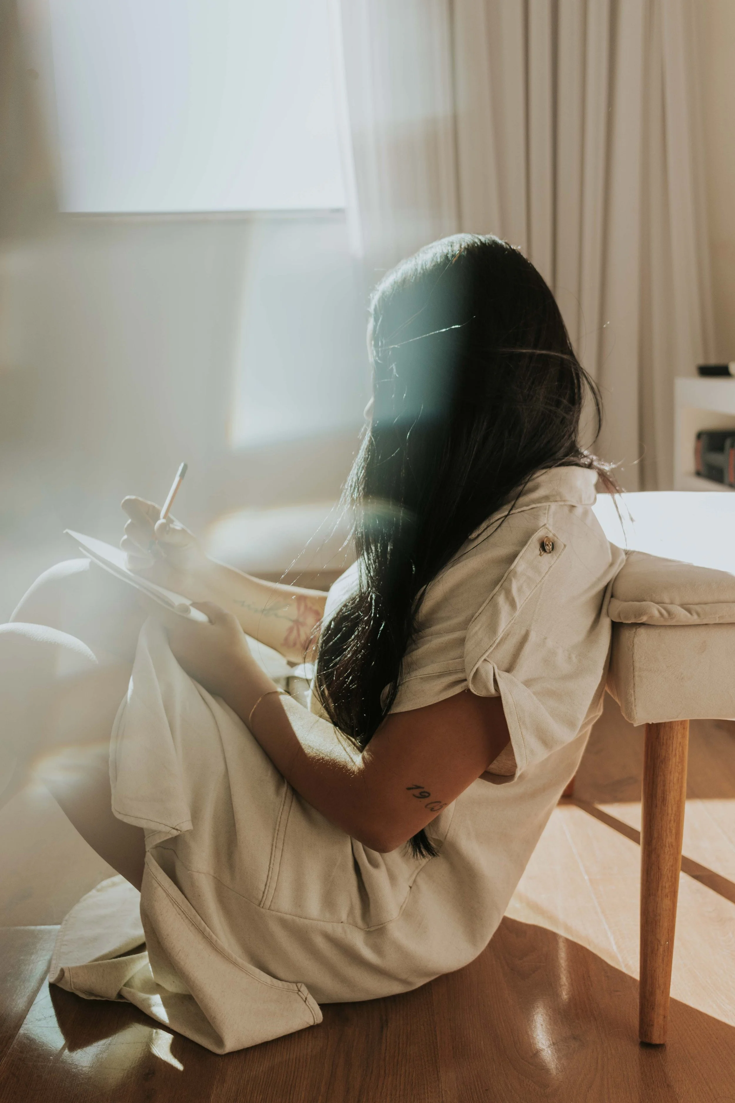 A woman sitting on the floor by a sunlit window, journaling quietly.