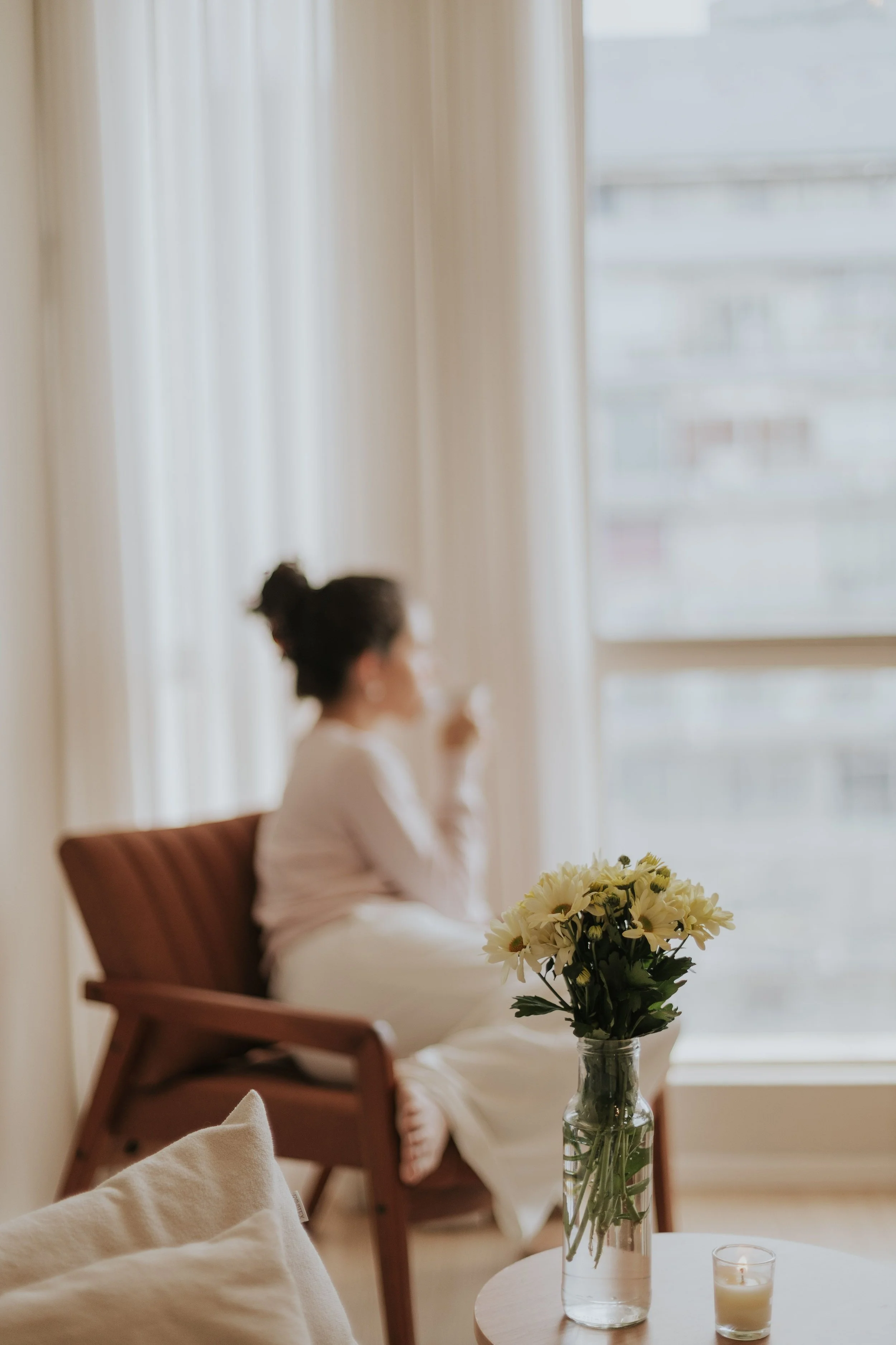A woman sitting in a chair by a window, holding a cup and looking out, with flowers on a nearby table.