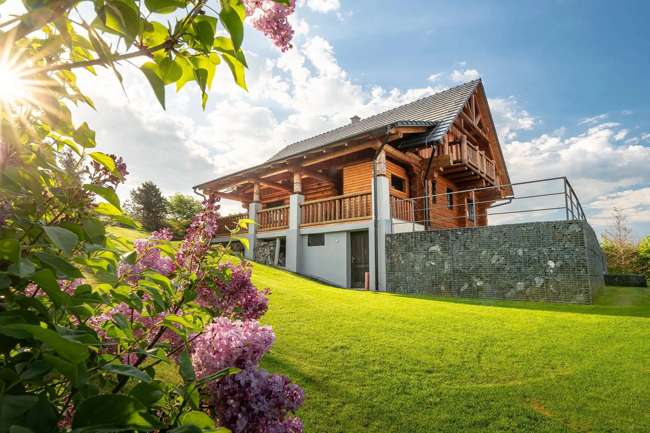 A wooden house with a balcony, set in a lush green yard. The house has a gray tiled roof and is surrounded by vibrant pink and purple flowers and green foliage. The sky above is blue with some clouds, and sunlight is shining from the left side of the