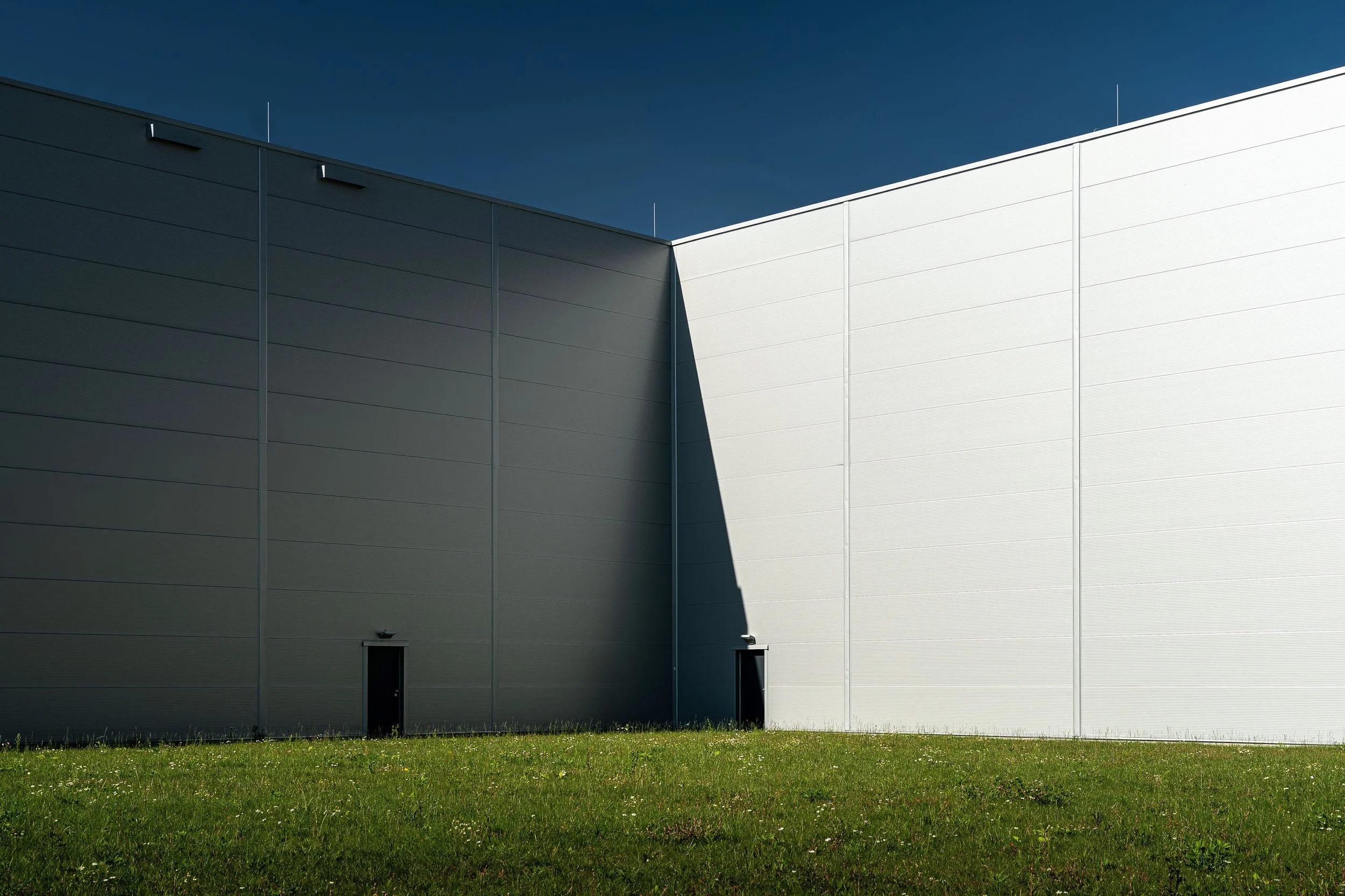 A modern building with gray and white panels on exterior walls, a door near the bottom, and a grassy area in the foreground. The sky above is clear and blue.