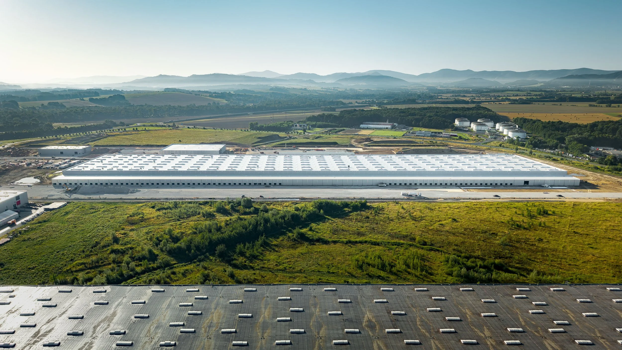 Aerial view of a large industrial building with a parking lot, surrounded by greenery and fields, with rolling hills in the background.