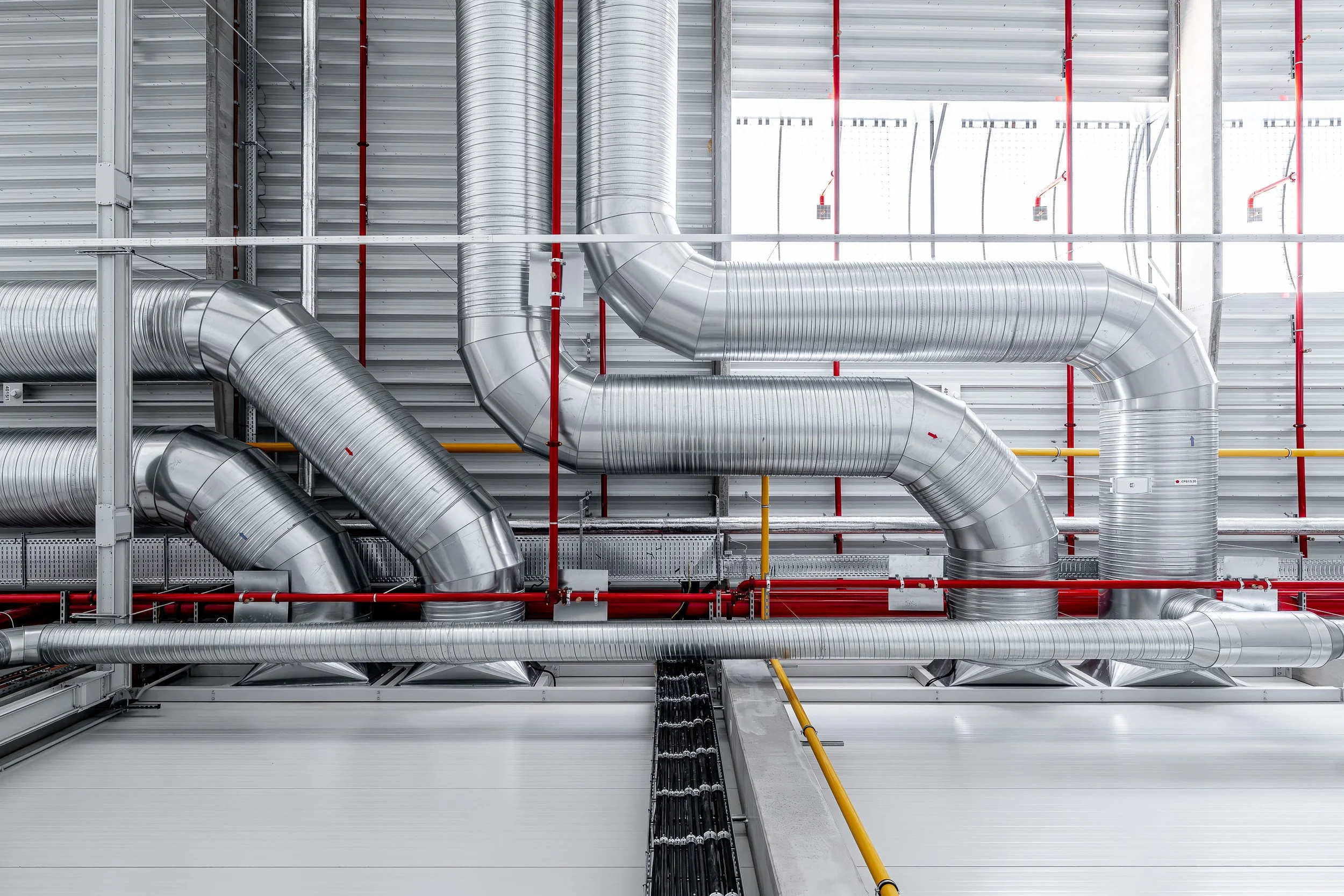Industrial ductwork system with large silver air ducts, red pipes, yellow piping, and support structures mounted on a metal wall ceiling.