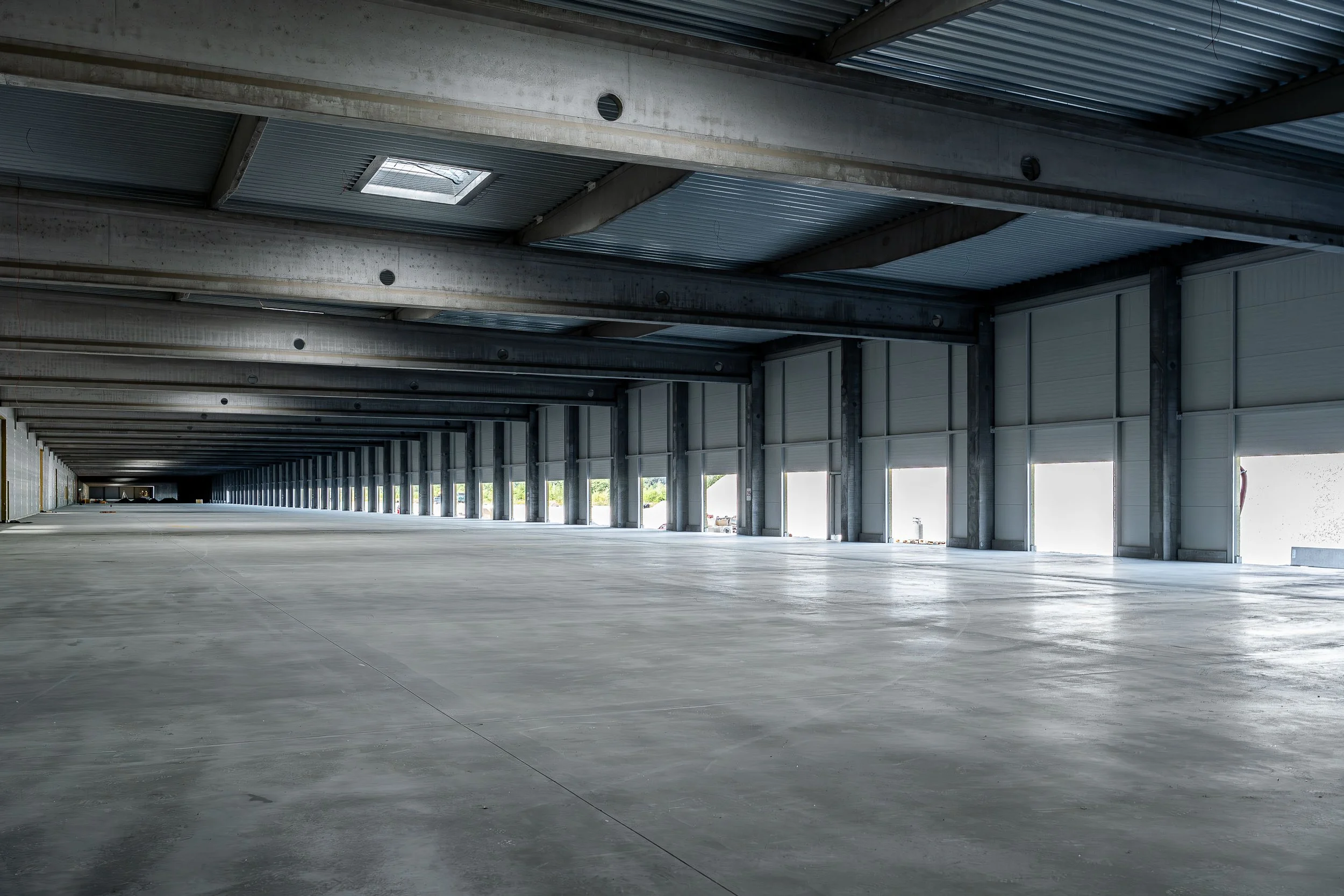 Interior of a large, empty industrial warehouse with a high ceiling, metal beams, and large open garage doors letting in natural light.