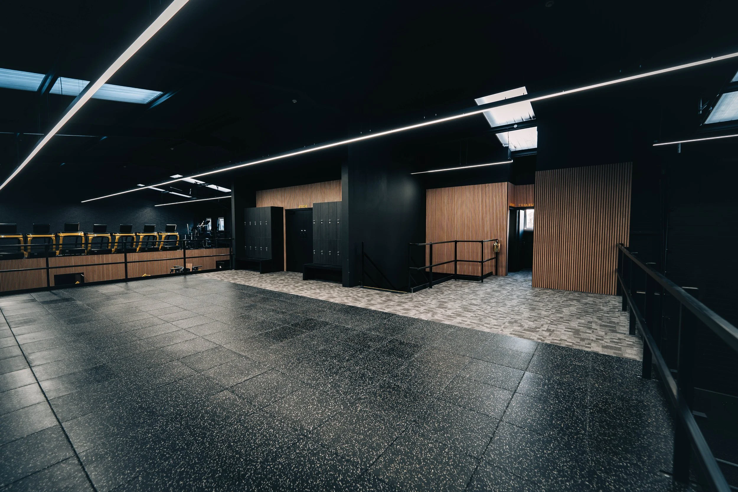 Empty modern indoor space with black walls, wooden accents, lockers, and a row of chairs in the background.