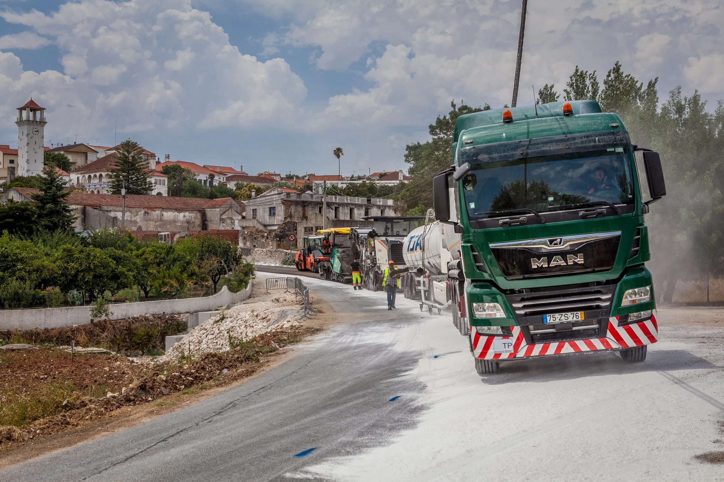 Imagem aérea de uma área agrícola com caminho de terra em construção, cercado por plantações de árvores e árvores de oliveira, com um cenário de colinas ao fundo.