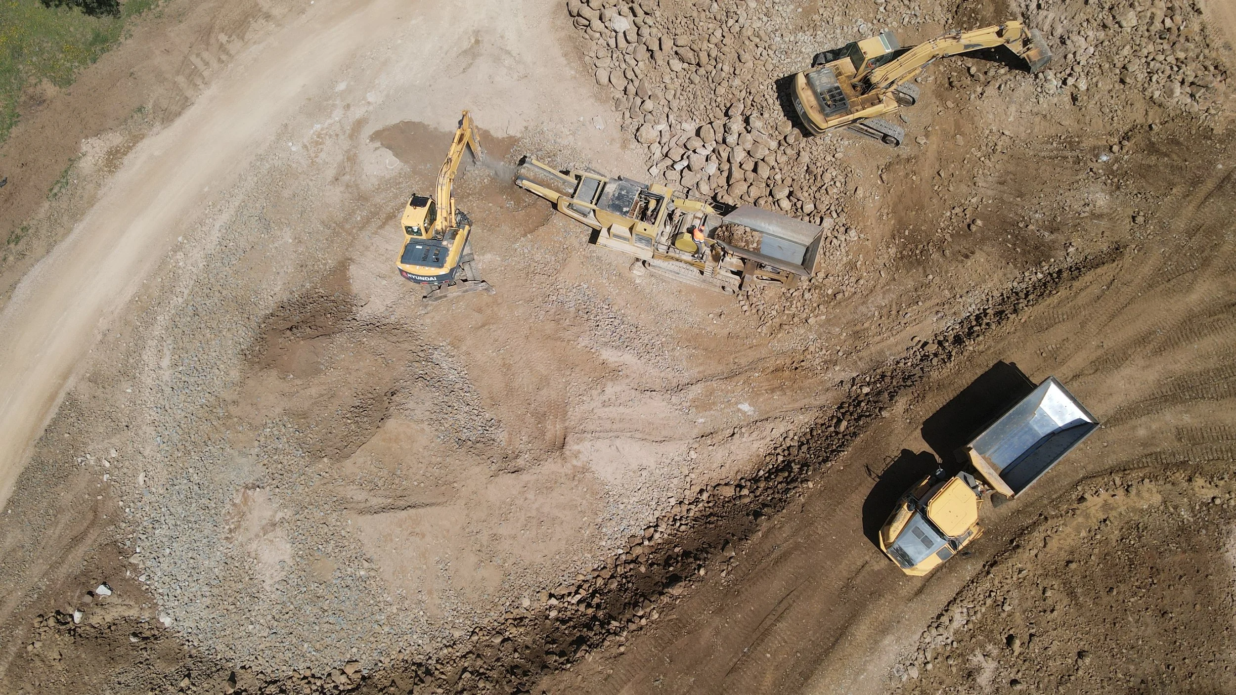 Homens colocando uma barreira de contenção de areia na orla de um corpo de água, com máquinas pesadas ao fundo e céu nublado.