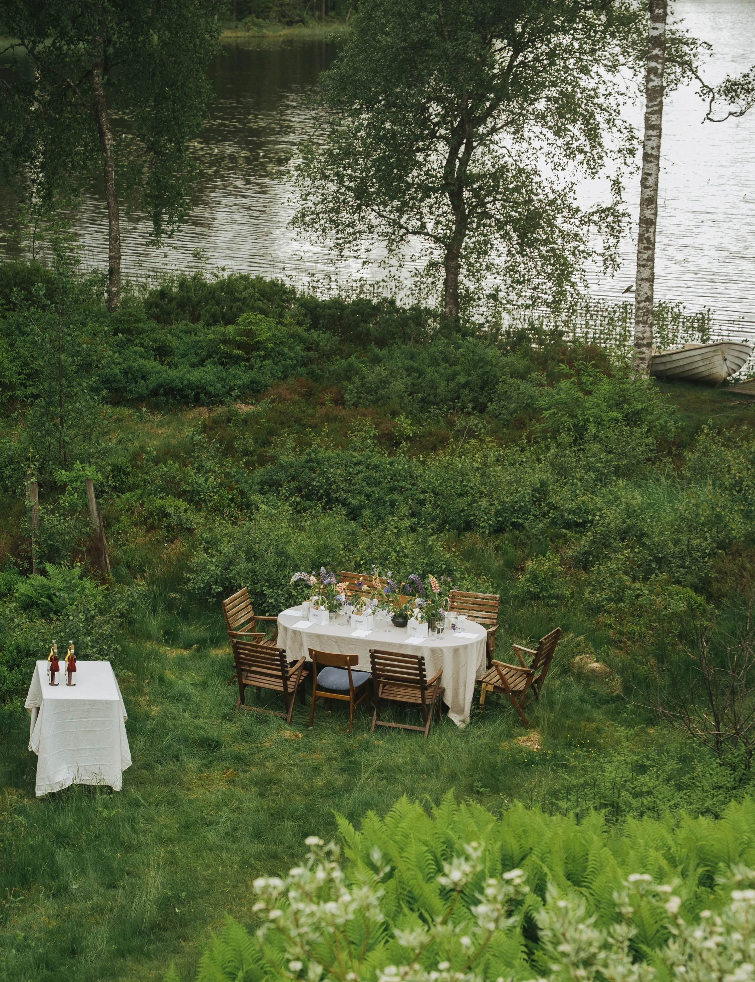 A menu on a table with a sketch of a table setting, next to a white plate with a glass vase containing green leaves, and a small glass of water. The menu includes aperitive, main, and dessert sections.