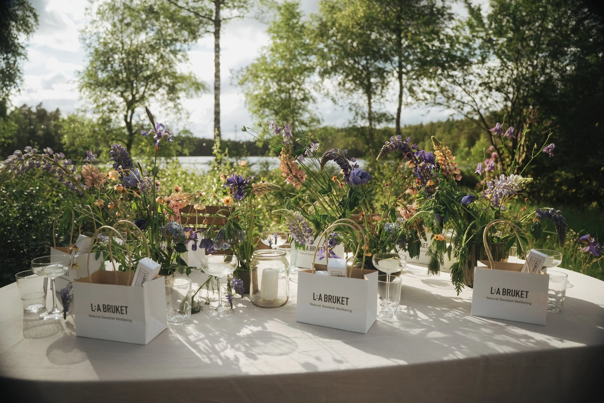 A dining table set outdoors with a goodie bag from "L:A BRUKET" placed on a plate, accompanied by a fork, knife, and spoon. The table features candles, glasses, and small vases with purple flowers and greenery, illuminated by warm sunlight.