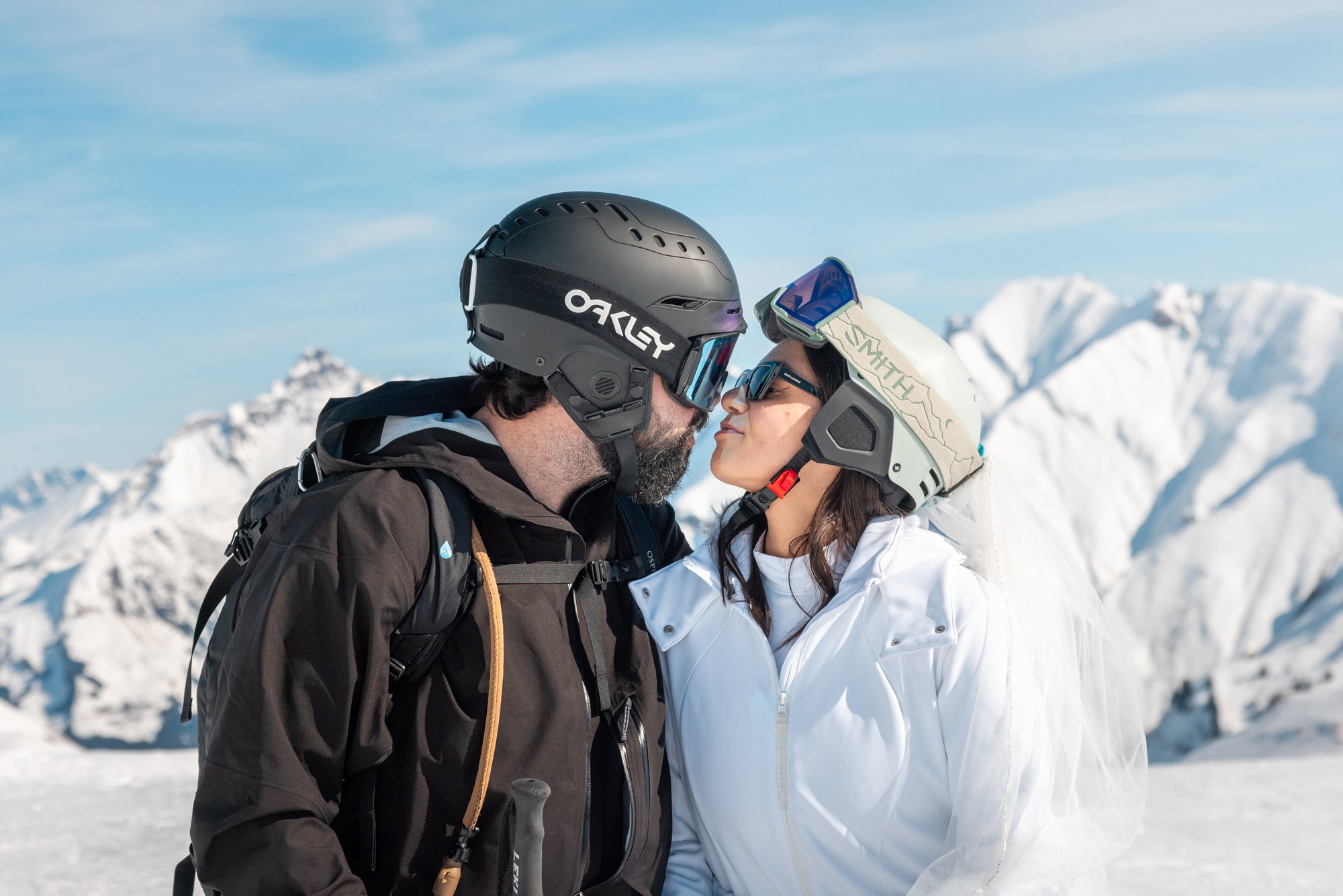 A couple in ski gear, touching foreheads, standing close on a snowy mountain with a clear blue sky and snow-covered peaks in the background.
